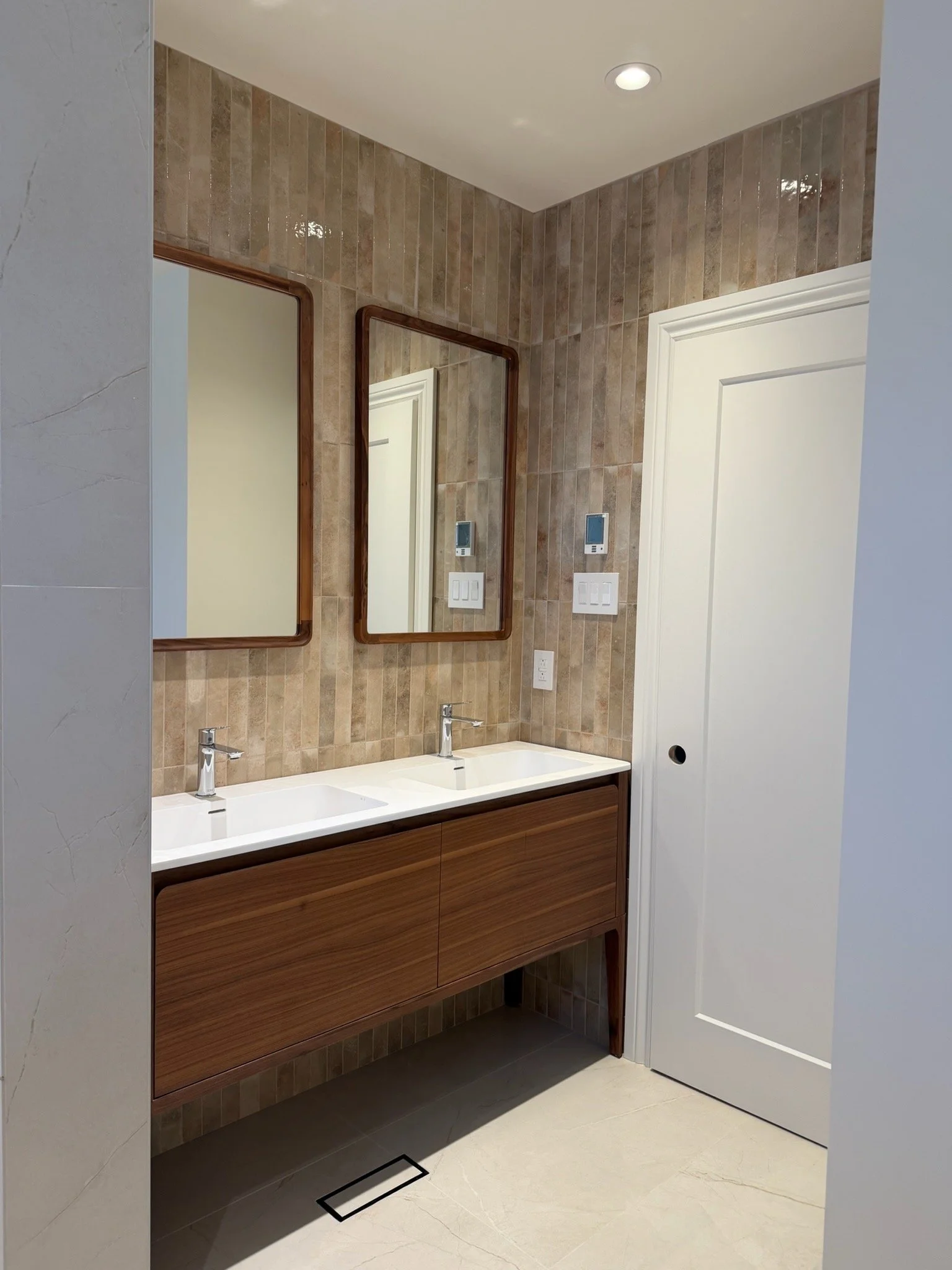 Bathroom with double sinks, vanity with wood finish, two mirrors, beige tile wall, and white door.