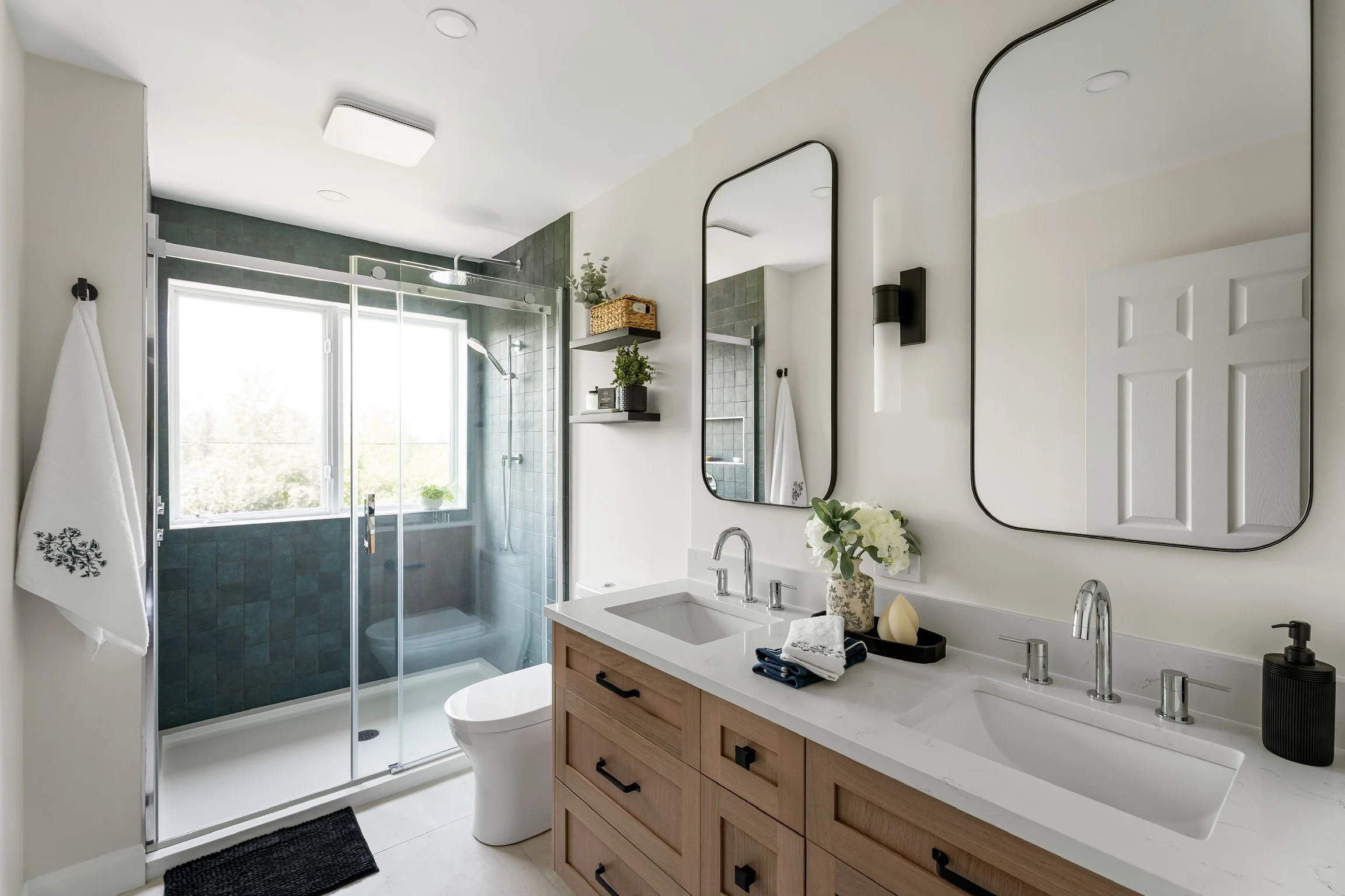 Modern bathroom with a double sink vanity, large mirrors, and a glass-enclosed shower with dark tiles and a window. Decor includes towels, a small plant, and decorative objects.