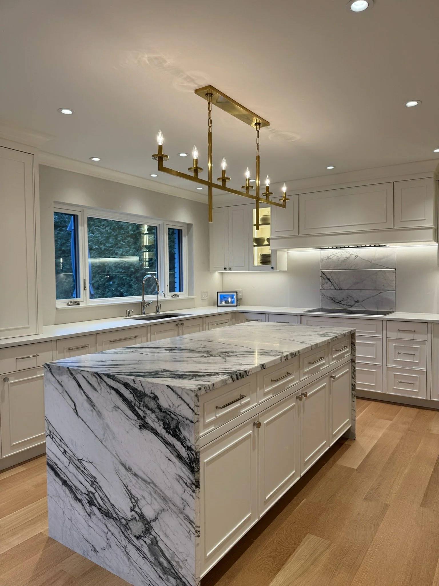 Modern kitchen with white cabinets, marble island, large window, brass chandelier, and recessed ceiling lights.