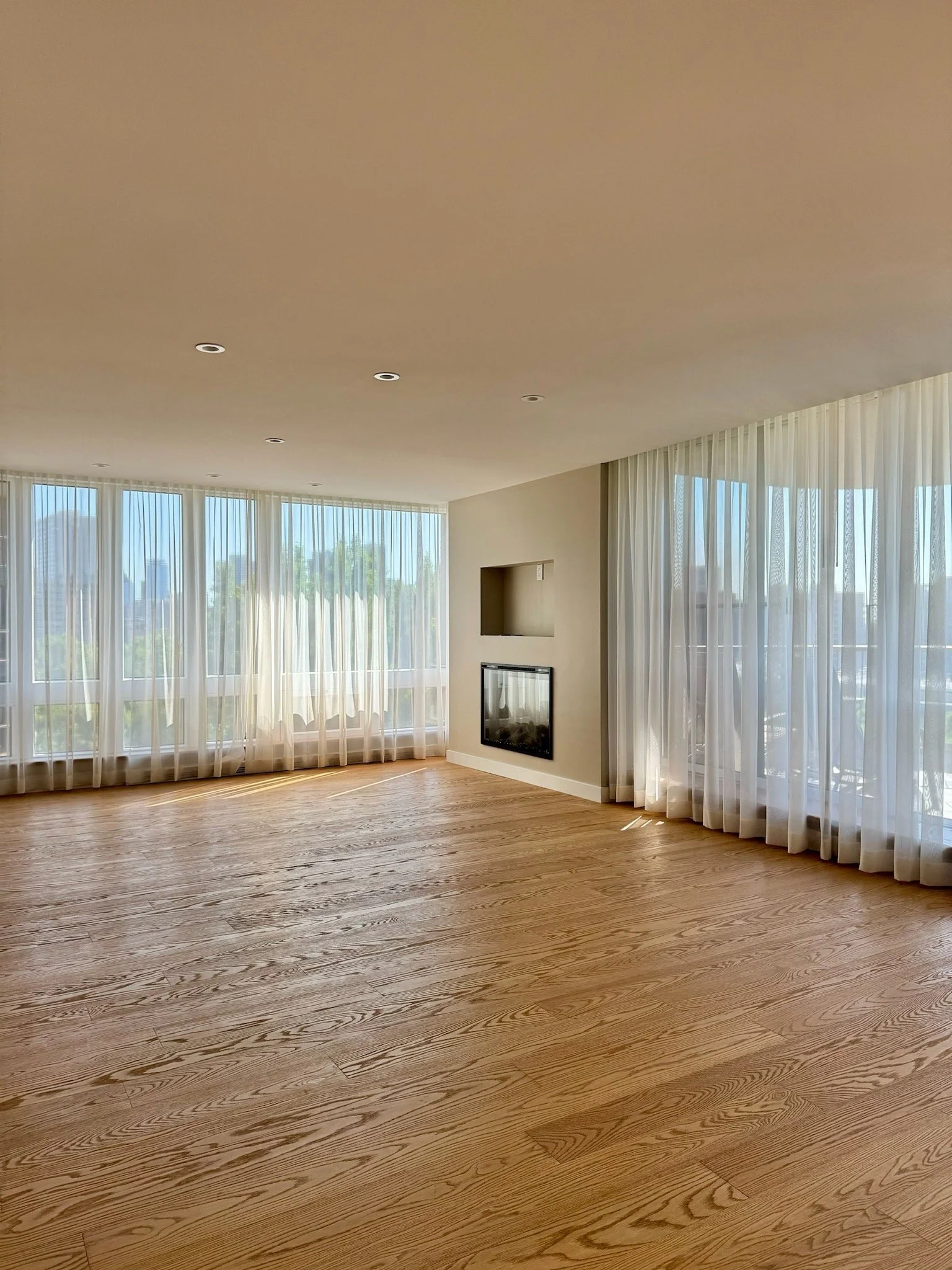 A bright living room with large windows covered by sheer white curtains, hardwood floors, a wall-mounted fireplace, and a recessed wall area.
