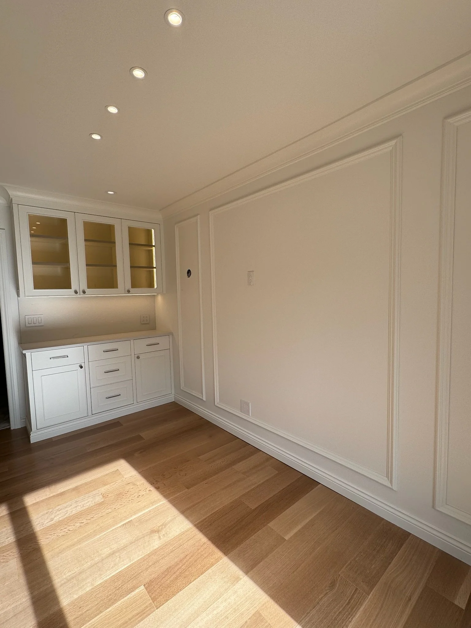 Interior view of a room with white cabinetry, crown molding, beige walls, and hardwood floors, illuminated by ceiling lights.