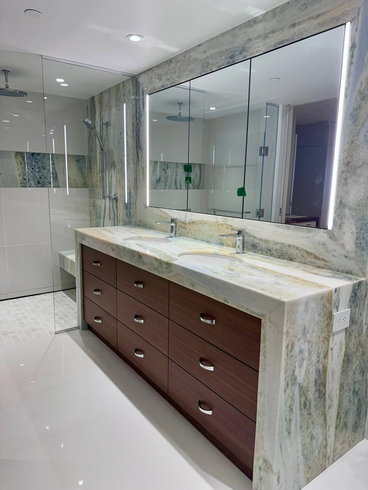Modern bathroom with a double sink vanity, marble countertop, large mirror with integrated LED lighting, glass shower enclosure with rainfall showerhead, white tiled floor, and minimalist design.
