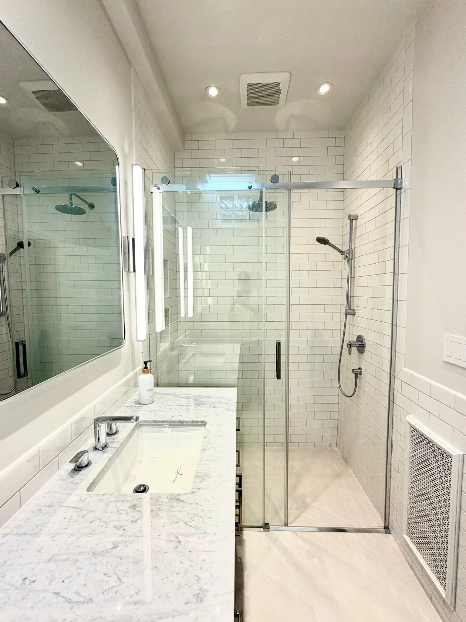 Modern bathroom with a white marble countertop, white subway tile walls, a glass shower stall with two showerheads, a large mirror, and a white vent on the wall.