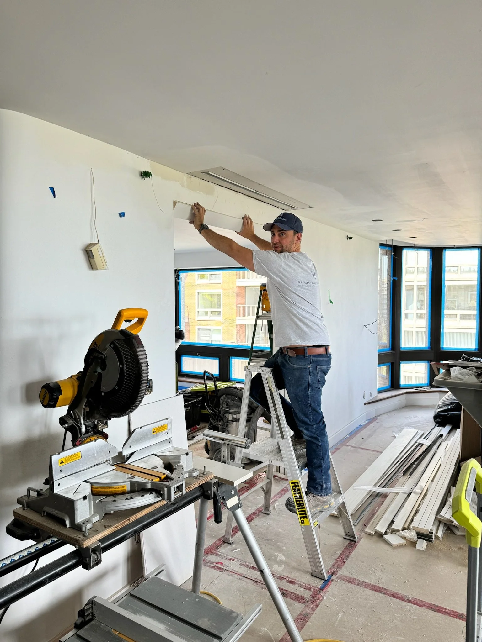 A man working on construction inside a room, standing on a ladder, surrounded by tools and materials.