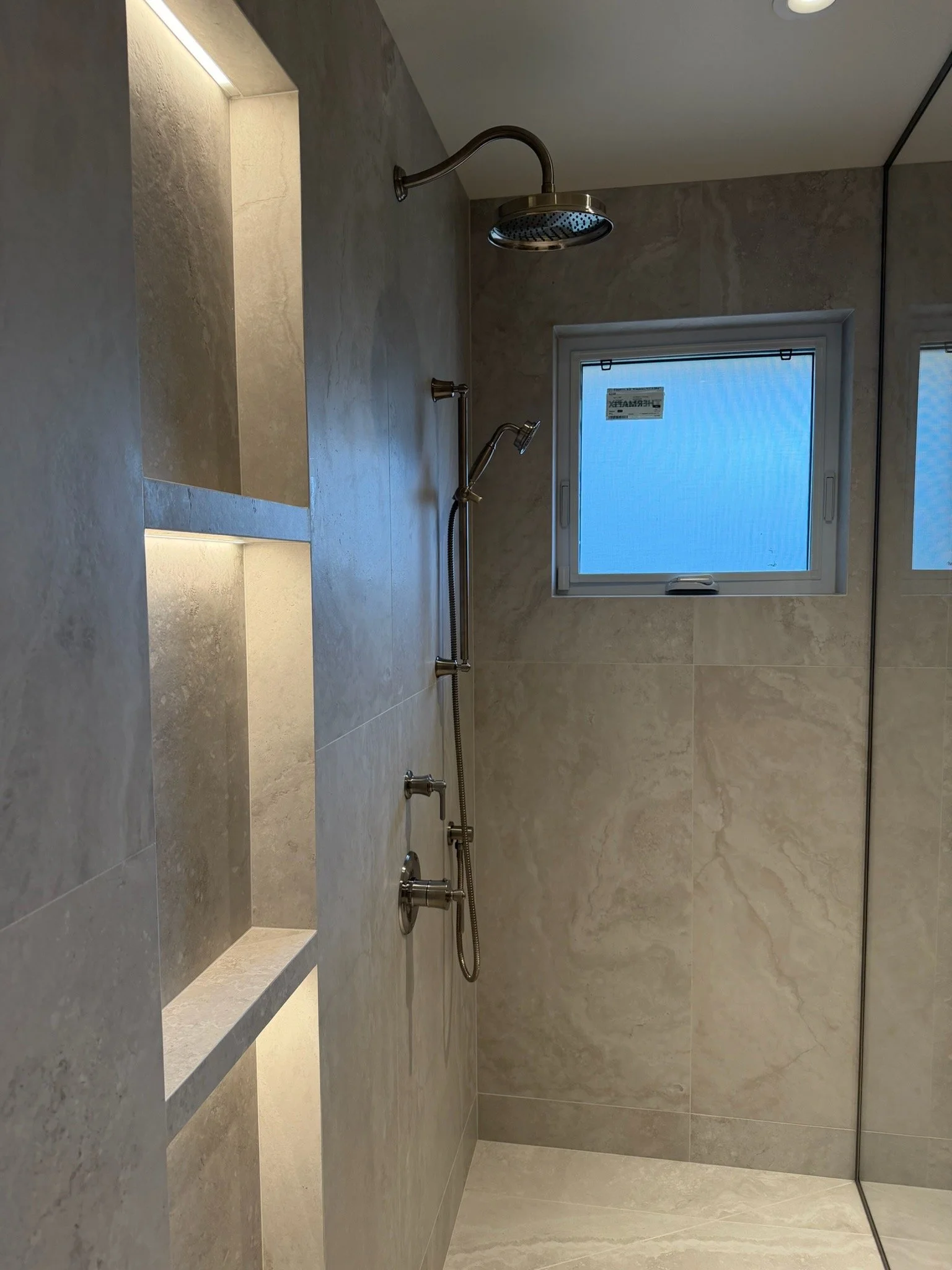 A modern shower with a built-in bench, grey tiled walls, a fixed window, a large rainfall showerhead, a handheld showerhead, and a mirror wall.