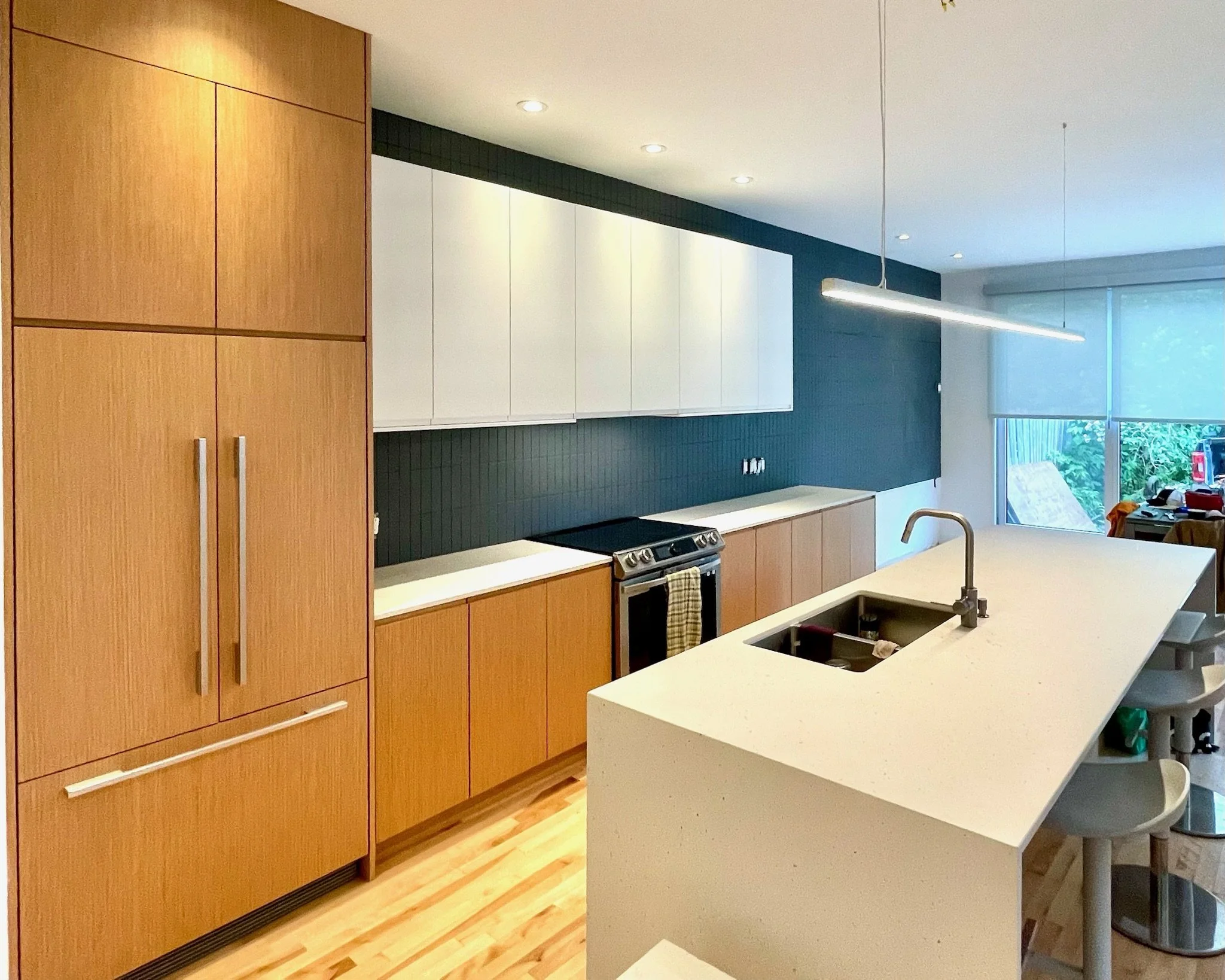 Modern kitchen with wooden cabinets, a white island with a built-in sink, black backsplash, and nearby large window with a view of outdoors.