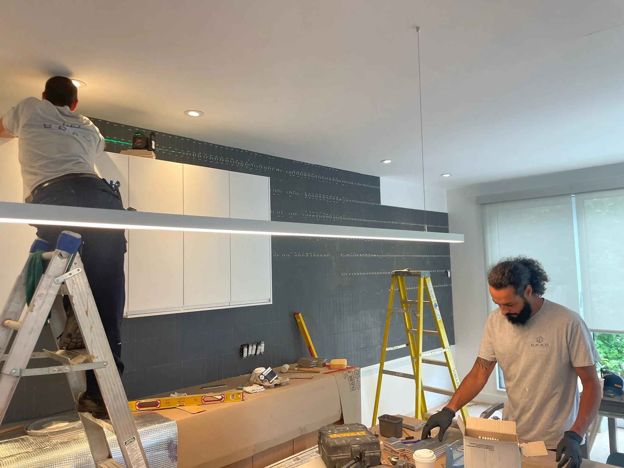 Two workers installing cabinetry in a kitchen. One is on a ladder working on the upper cabinets, the other is working on the lower countertop. Office supplies and tools are scattered on the counter. The room has large windows and a modern design.