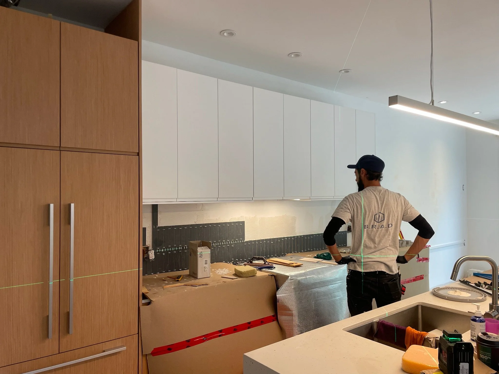 A person working in a modern kitchen under construction or renovation, with tools and materials on the countertop and a large wooden cabinet on the left side.