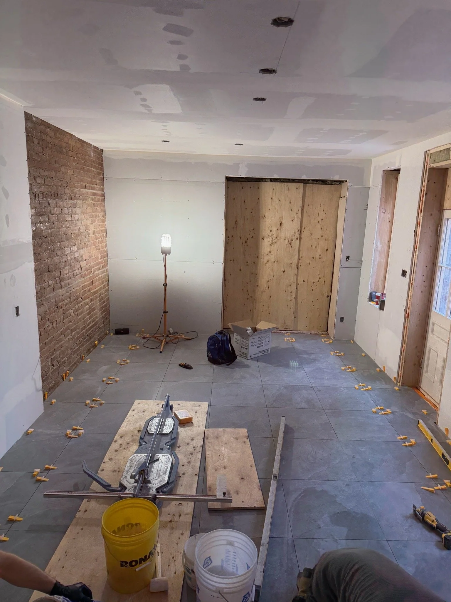 A room under construction with drywall being installed, a brick accent wall on the left, and various construction materials and tools scattered on the floor, including buckets, a miter saw, and a work table.