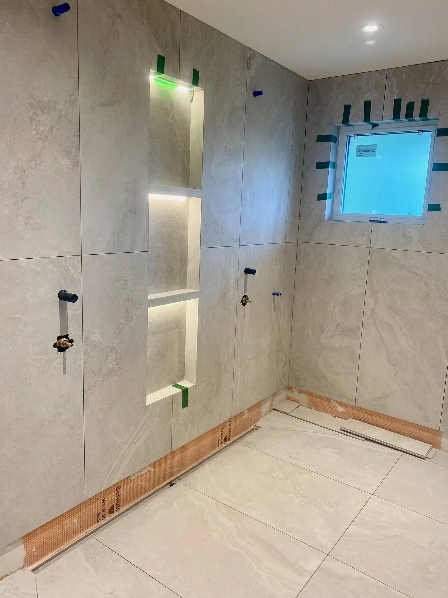 Bathroom under construction with large beige marble tiles, installed mirror, and a window with masking tape.