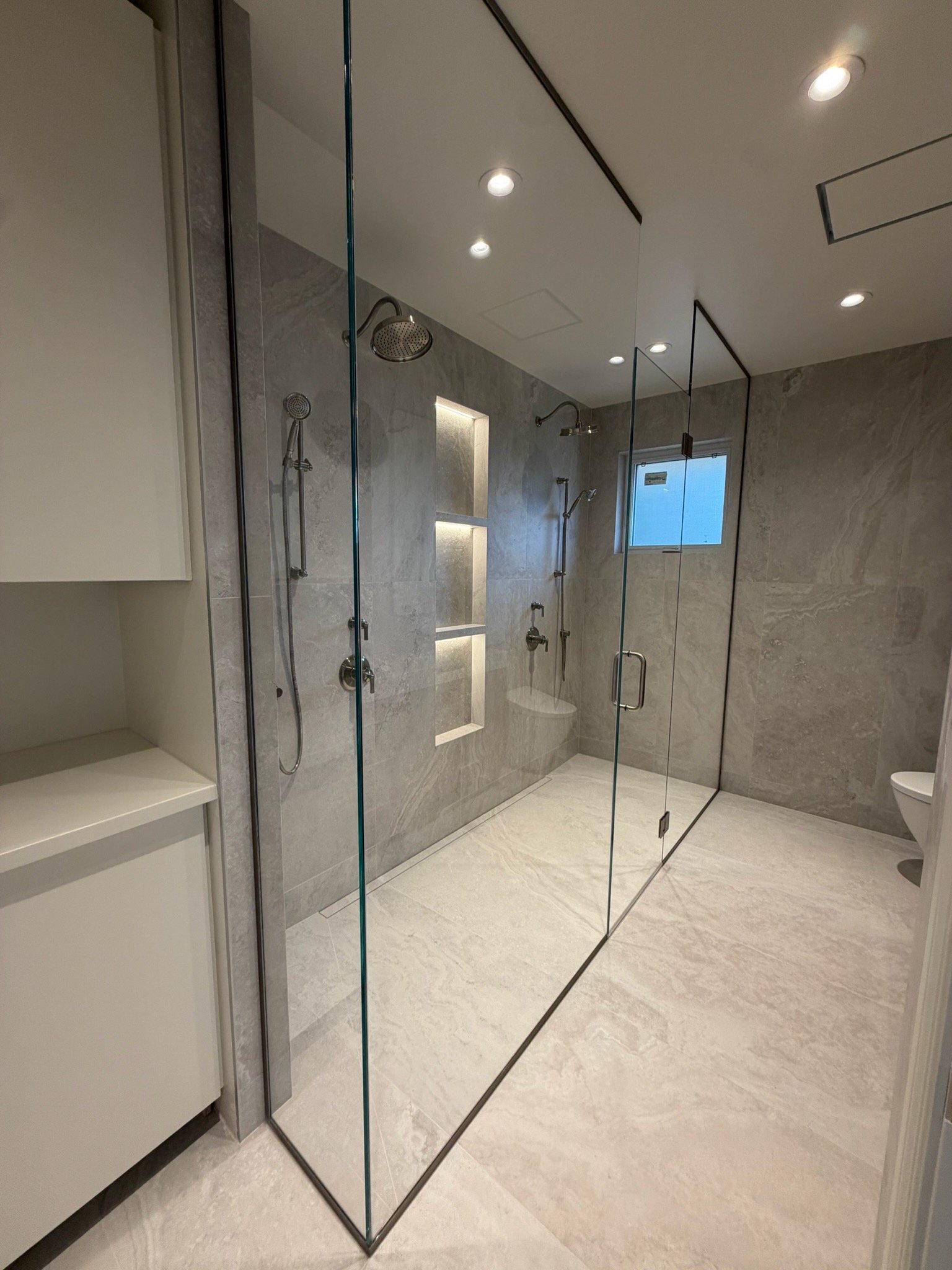 Modern walk-in shower with glass enclosure, multiple shower heads, built-in shelves with lighting, and beige marble walls and floor.