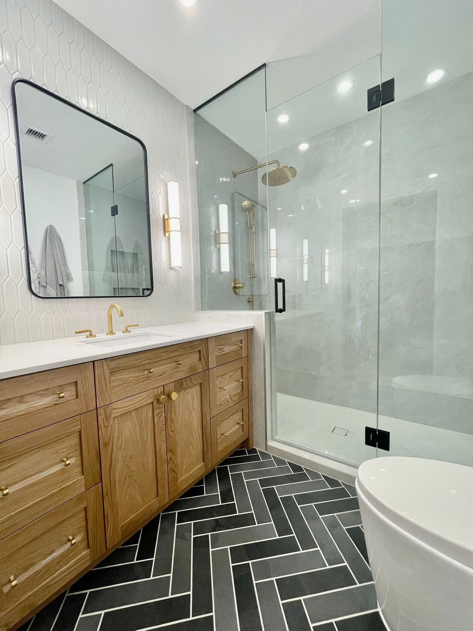 Modern bathroom featuring a wooden vanity with gold hardware, black and white herringbone floor tiles, a mirror, and a glass-enclosed shower with gold fixtures and a rainfall showerhead.