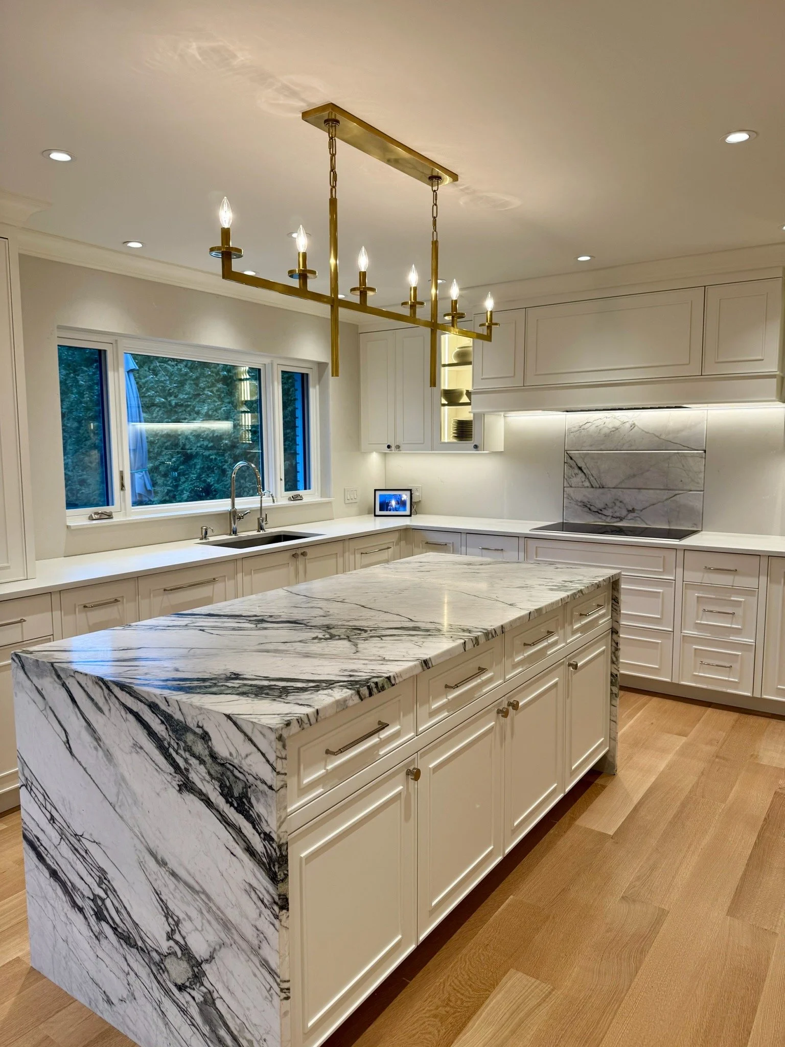 Modern kitchen with white cabinets, a marble island, a window view of trees, and a brass chandelier with candle-like light bulbs.