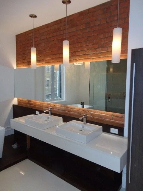 Modern bathroom with two white vessel sinks, large mirrors, three hanging pendant lights, exposed red brick wall, and a window.