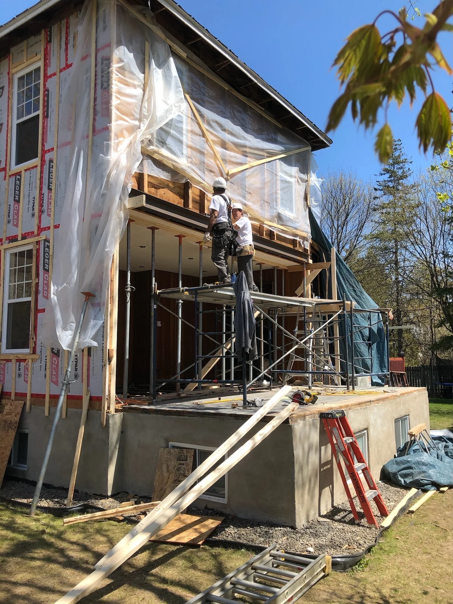 Construction workers on scaffolding building onto a house, with wood framing and plastic sheeting, on a sunny day with trees in the background.