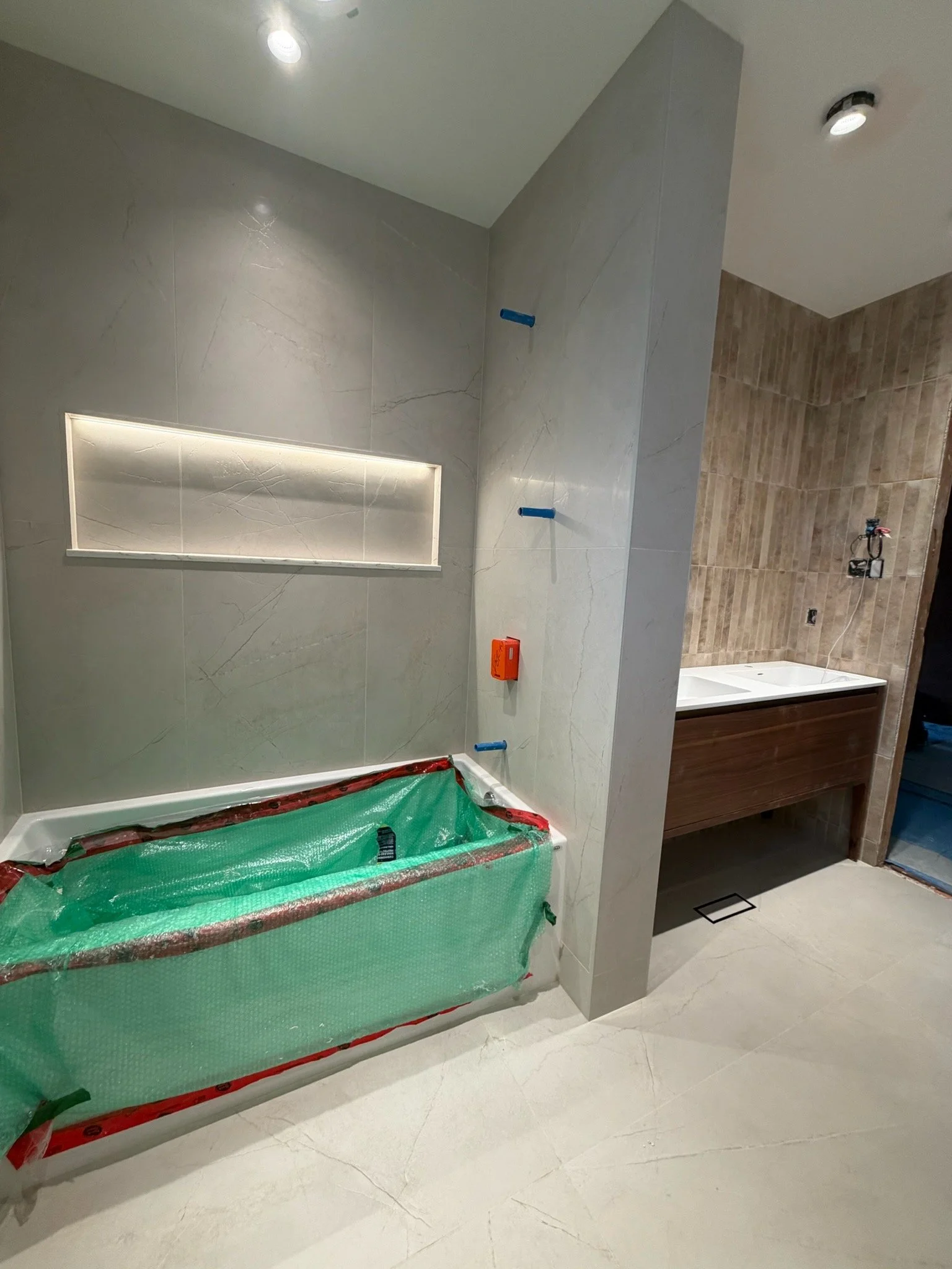 An unfinished bathroom with tiled walls and floor. A bathtub is wrapped in green protective plastic, and there are blue wall anchors on the wall. The bathroom has a small window with built-in lighting, and a wall-mounted vanity with a sink on the rig