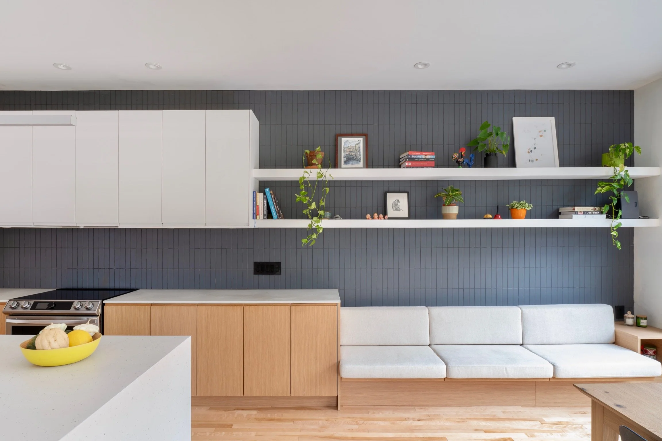 Modern kitchen with white and wood cabinetry, a white sofa, and a dark textured accent wall with floating shelves decorated with plants, books, and framed artwork.