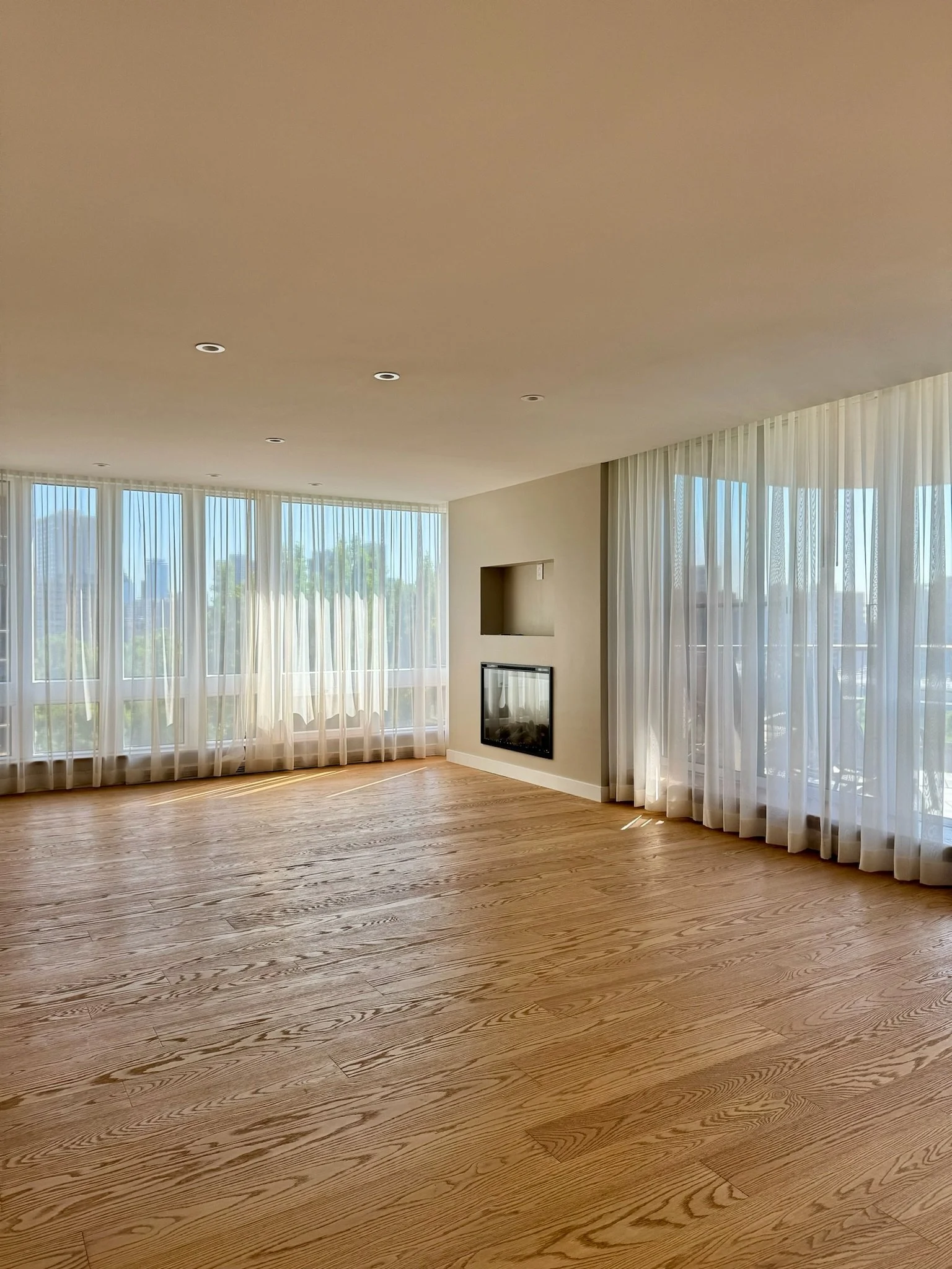 A spacious living room with large windows covered with sheer white curtains, hardwood flooring, and a wall-mounted fireplace with a niche above it, in a modern home.