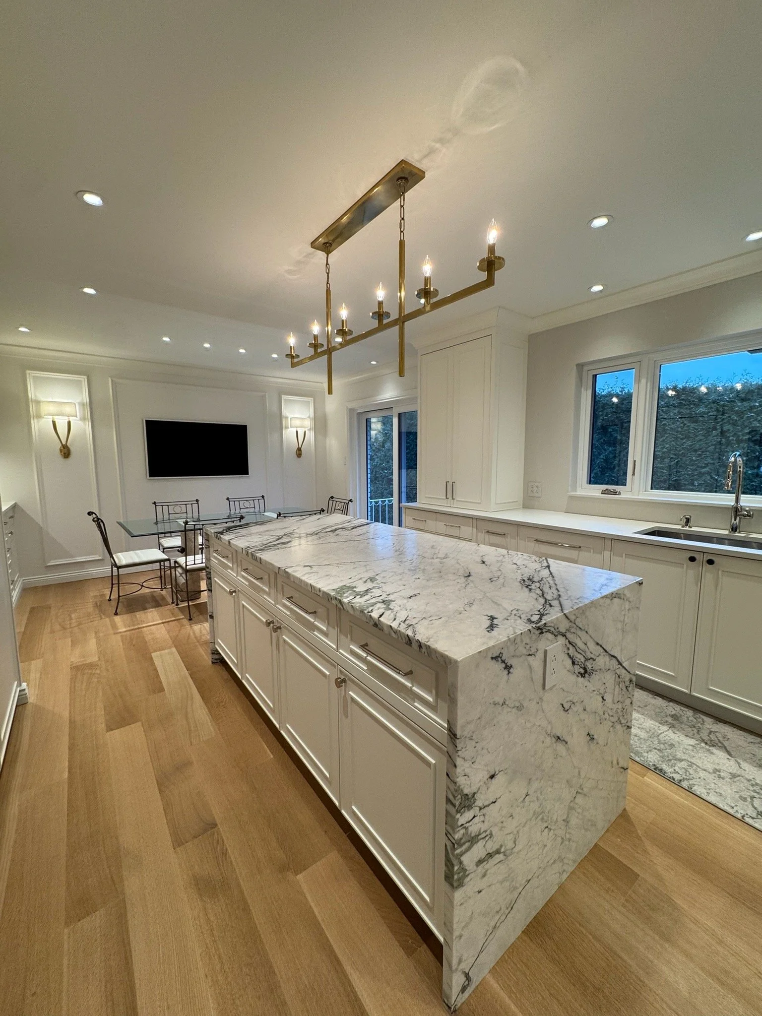 Modern kitchen with white cabinets, marble island, wooden floor, and a large window showing trees outside.