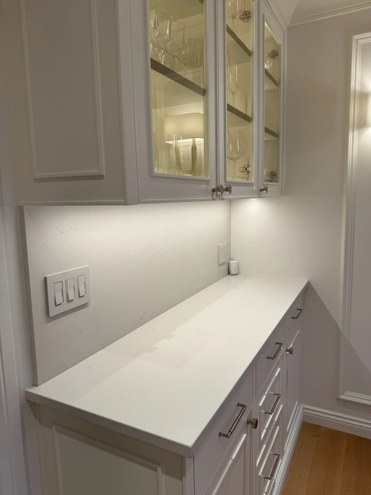A white kitchen cabinet with glass doors displaying wine glasses inside. Below, a white countertop with multiple drawers and a cabinet. Light switches and an outlet are visible on the wall.