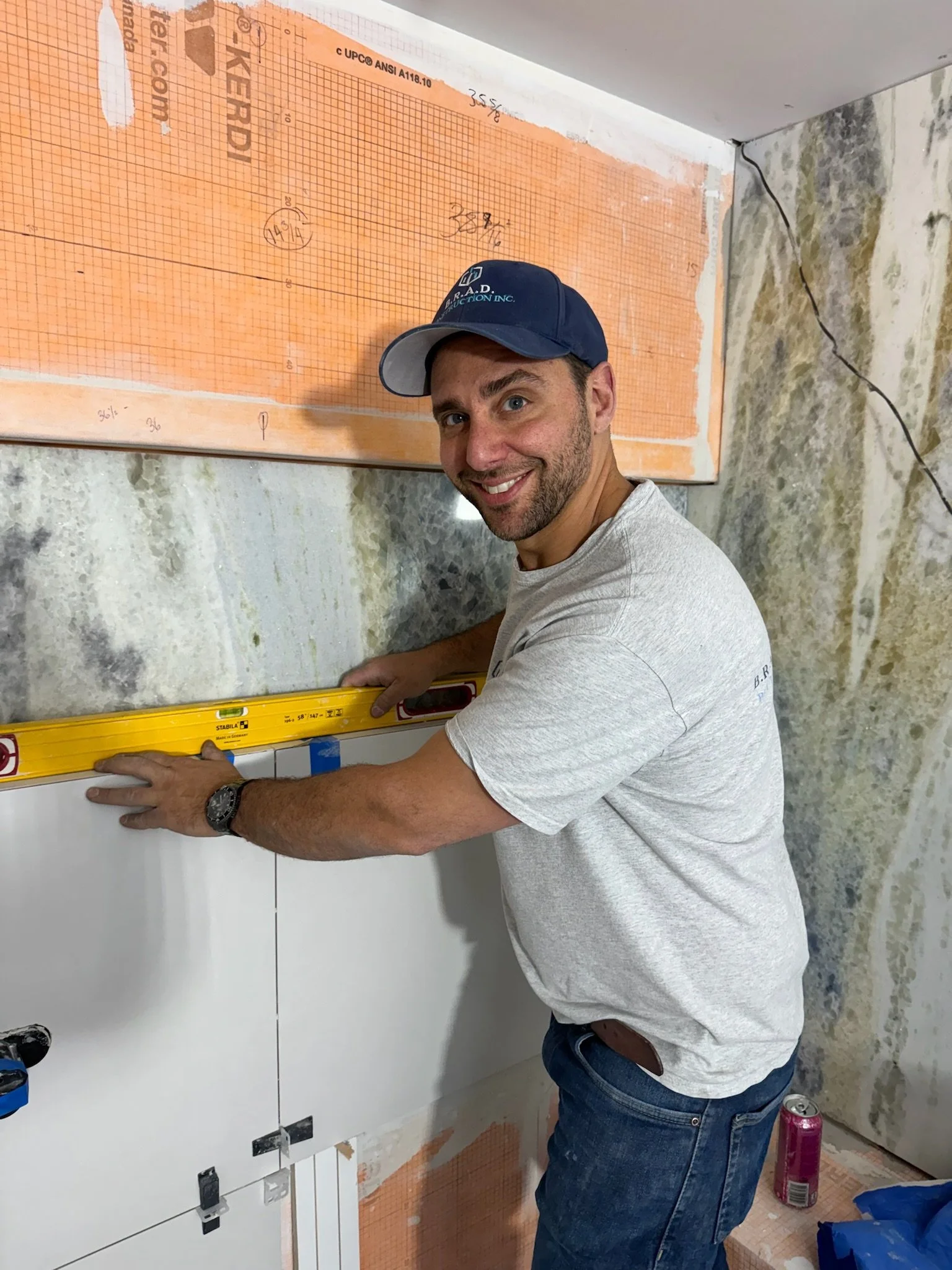 A man in a gray t-shirt and blue jeans is using a yellow level tool on a white tile wall. He wears a navy cap with text and a wristwatch, smiling at the camera. The wall behind him has a large orange grid pattern with some markings, and there are con