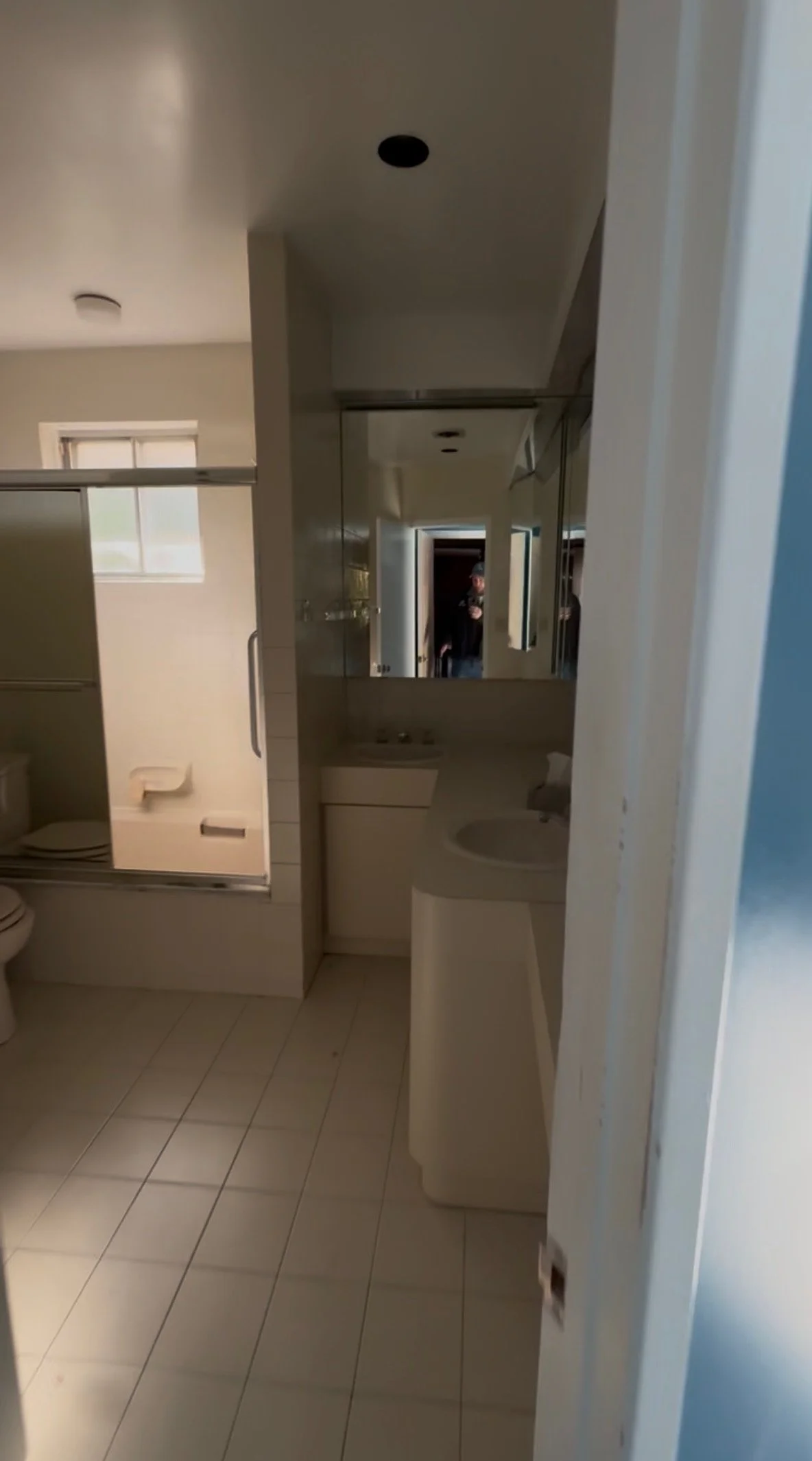 Bathroom with beige tiled floor, white fixtures, mirror, sink, and shower area; doorway and window visible.