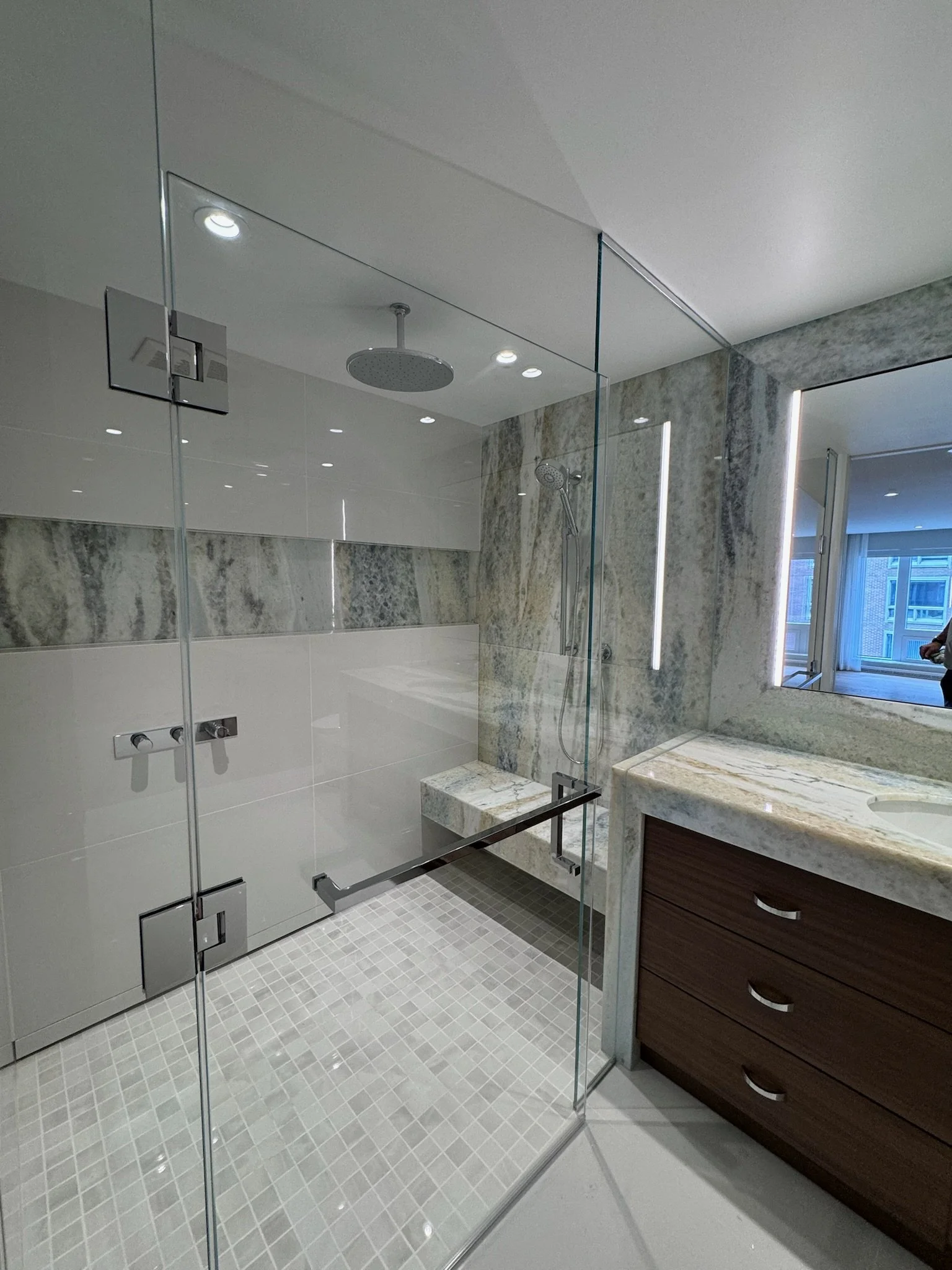 Modern bathroom with a glass shower enclosure, a rainfall showerhead, and a hand-held showerhead. The shower area has marble walls, a tiled floor, and a large mirror. A wooden vanity with drawers is visible, along with a window that allows natural li
