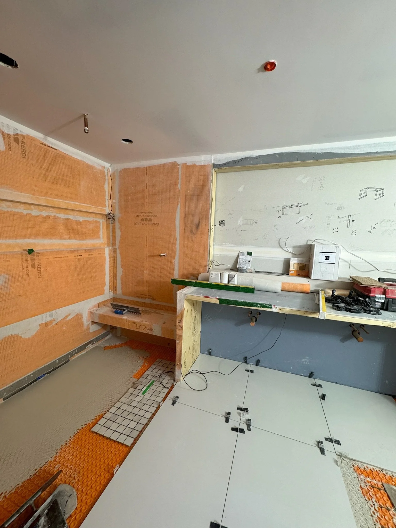 Interior view of a construction or renovation site with partially tiled and unfinished walls, tools, and materials scattered around, including a level, wires, and construction tape.