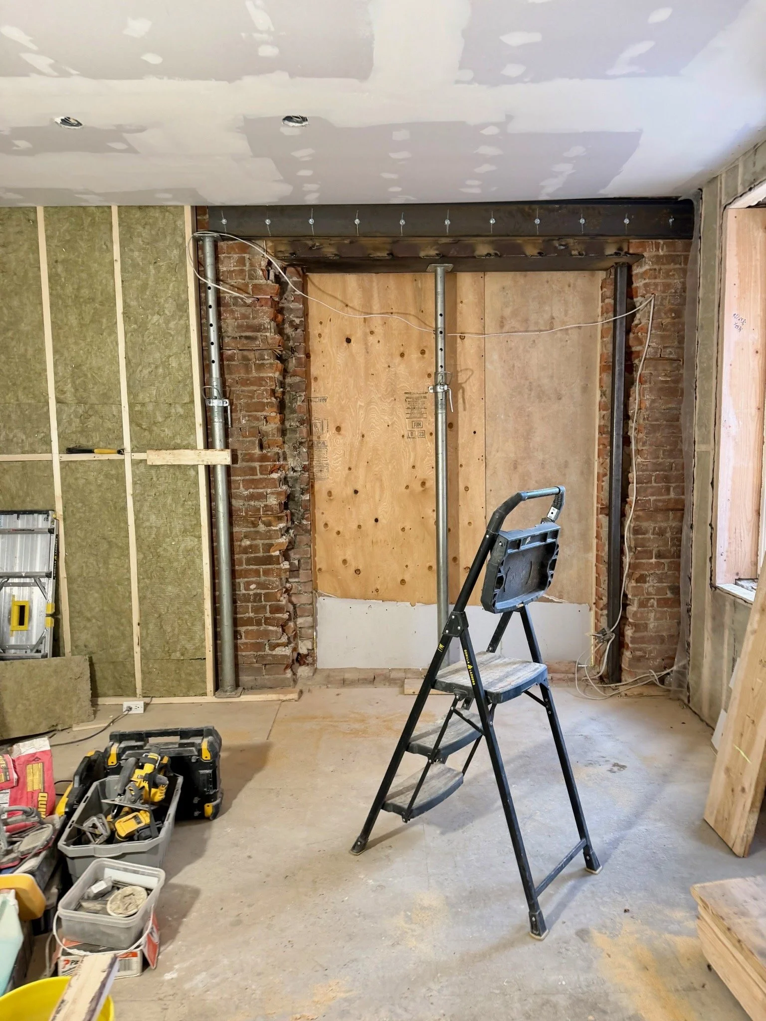 Interior of a construction site with exposed brick walls, drywall, construction tools, and a stepladder.