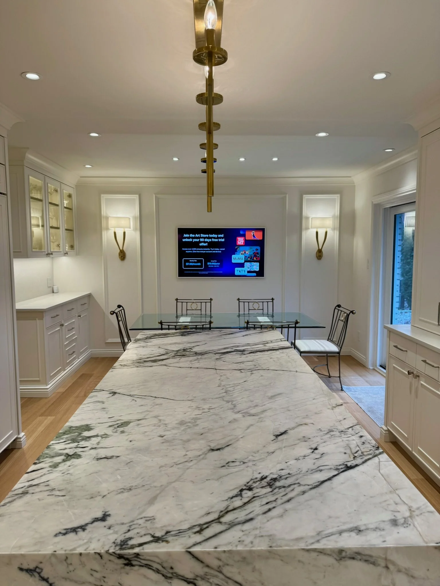 A modern dining room with a large white marble table with black veins, surrounded by four black metal chairs. The room has white walls with decorative molding, built-in white cabinets, wall sconces, and a flat-screen TV on the wall. There's a window 