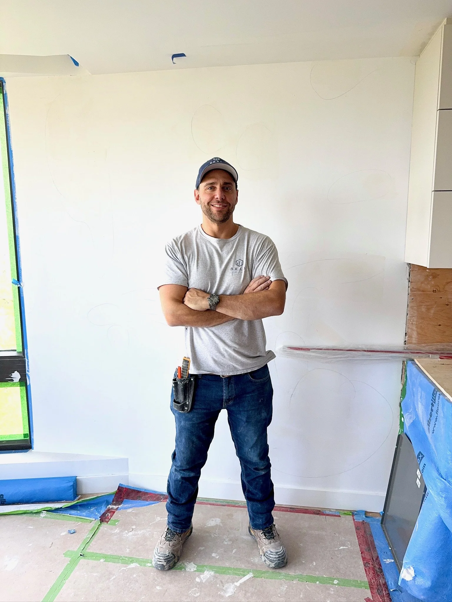A man with a beard wearing a gray t-shirt and a baseball cap stands with arms crossed in a room under renovation. The room has a partially finished drywall wall with faint circles drawn on it, paint supplies on the floor, and protective tape on the e