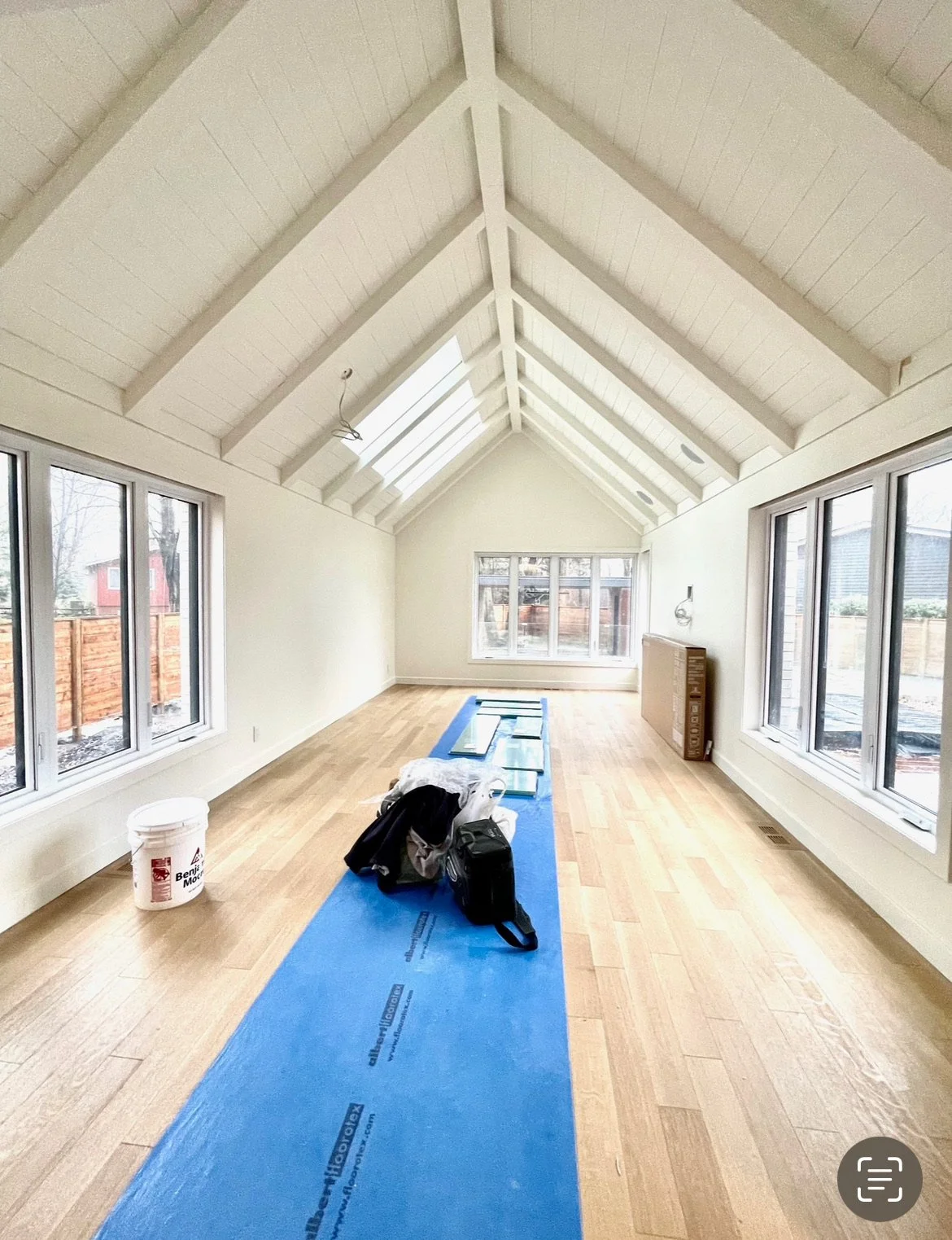 A bright, spacious room with a vaulted ceiling featuring white wooden beams and ceiling panels. Large windows on both sides let in natural light and offer views of a fenced backyard with some trees and neighboring houses. The floor is hardwood, and t