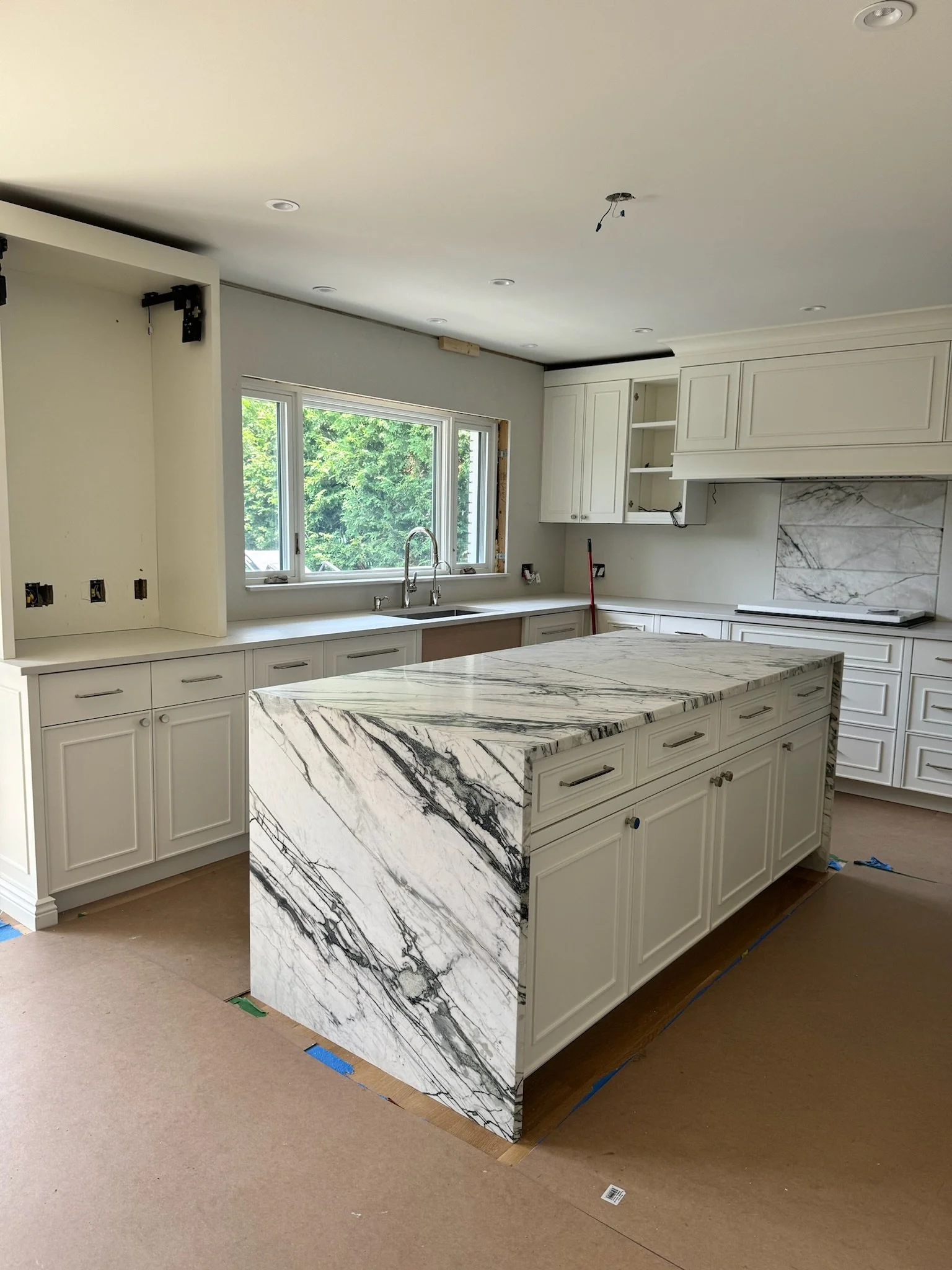 Kitchen with white cabinetry, marble island, window with green trees outside, under construction with missing light fixture and exposed wiring.