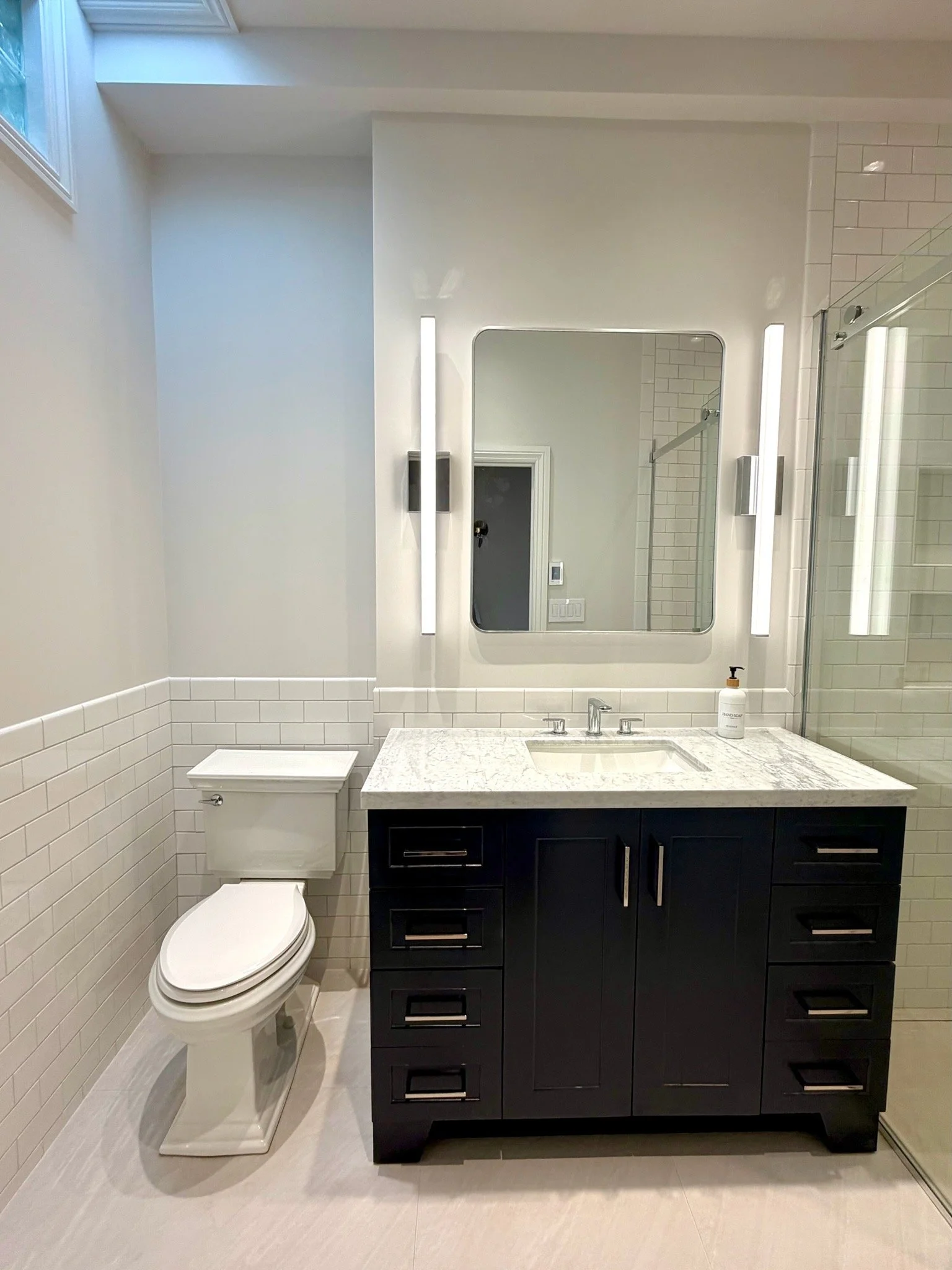 Modern bathroom with white subway tile walls, a white toilet, a black vanity with a white marble countertop, a rectangular mirror, and a shower with glass doors.
