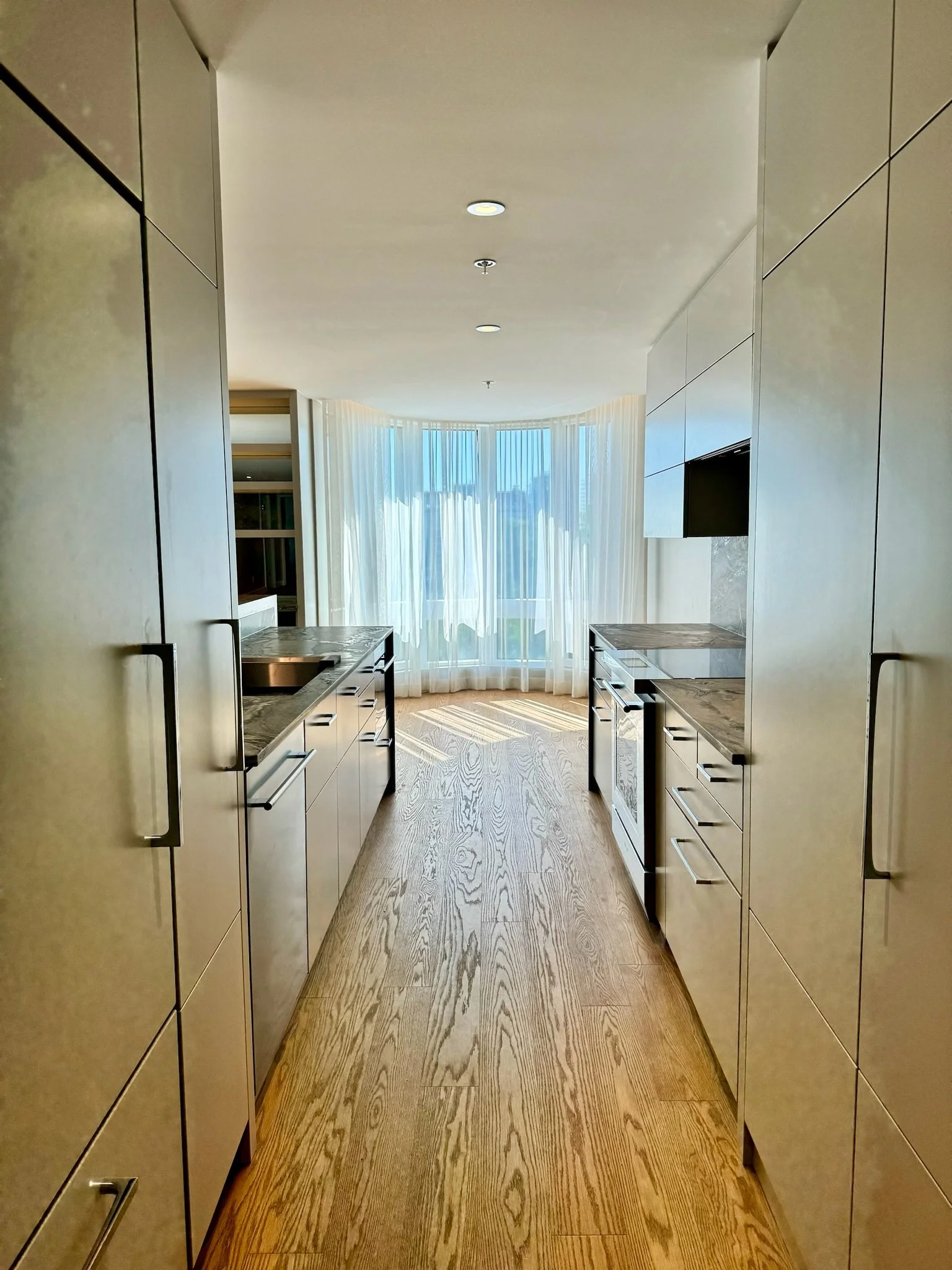 A modern, well-lit kitchen with beige cabinetry, marble countertops, and hardwood flooring. A large window with sheer curtains at the end of the kitchen allows natural light to fill the space.