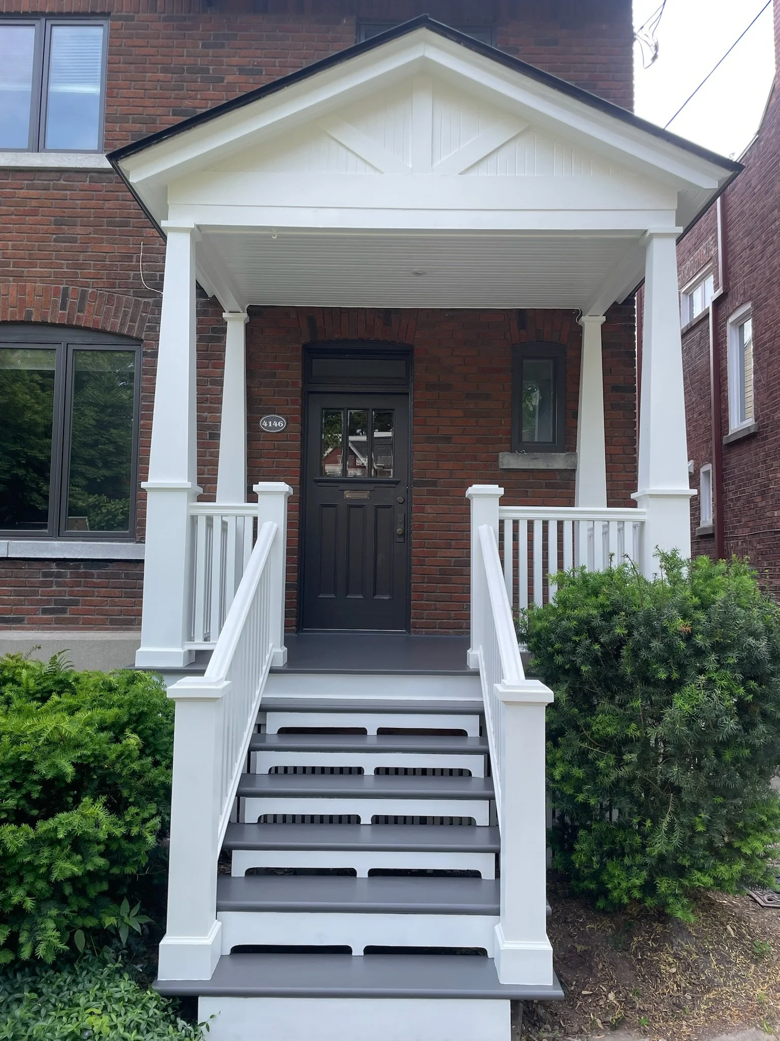 Front porch with black door, white railing, stairs, and brick house with a window.