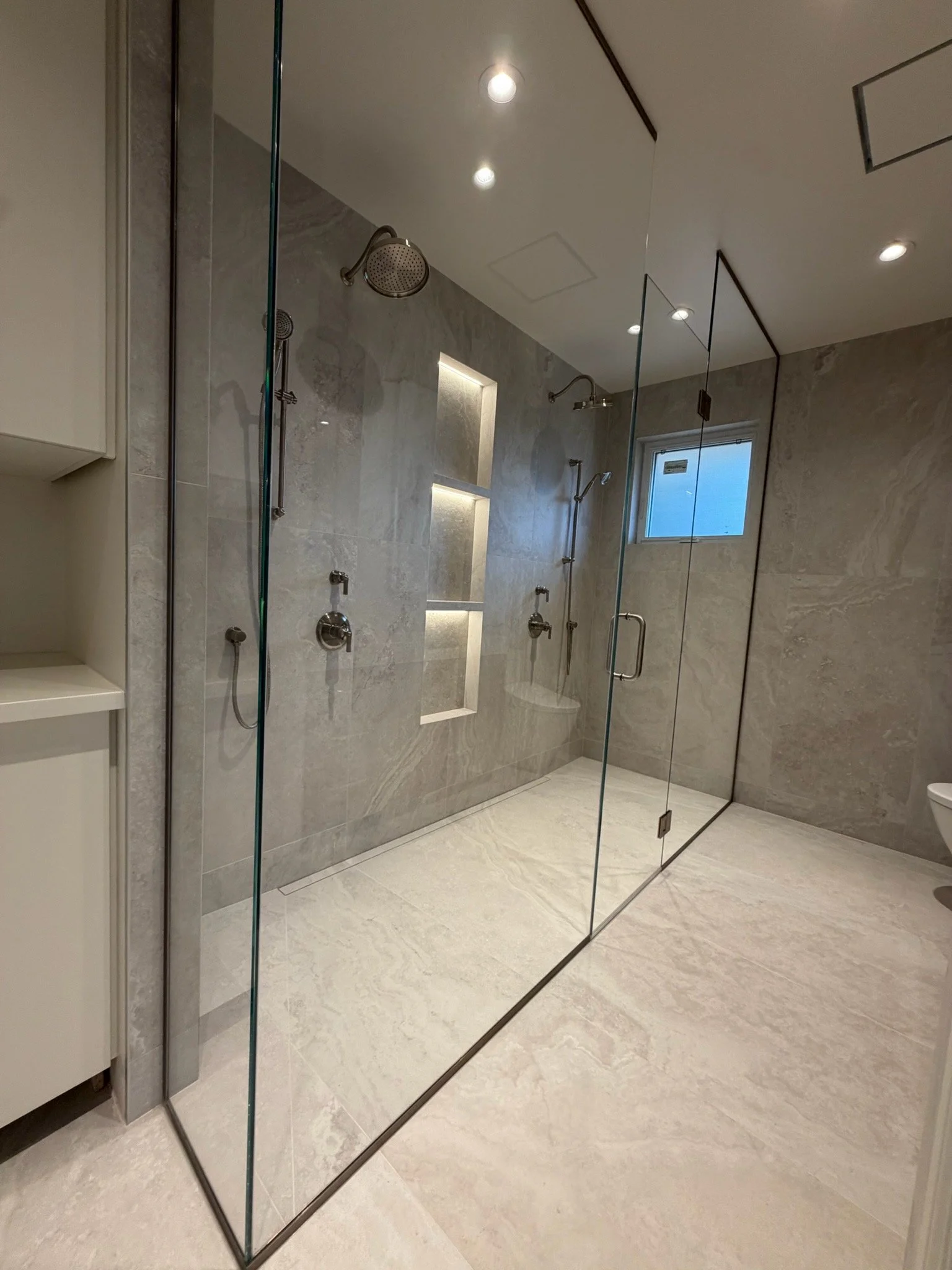 Modern bathroom with a large glass shower enclosure, marble walls, multiple shower heads, and built-in wall niches for toiletries.