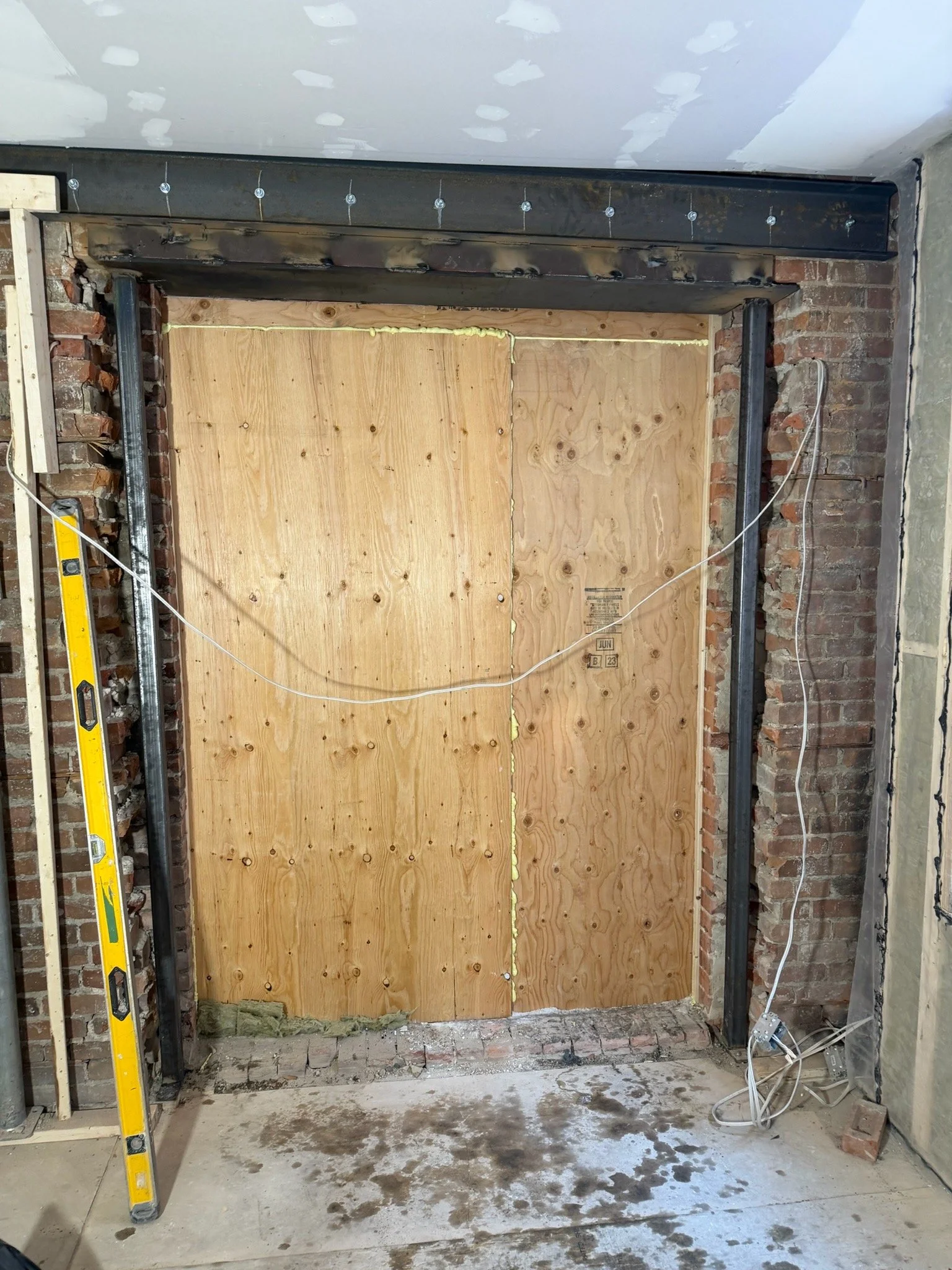 Construction site with brick wall, wooden panel, metal framing, electrical wiring, and a yellow ladder in an unfinished room.