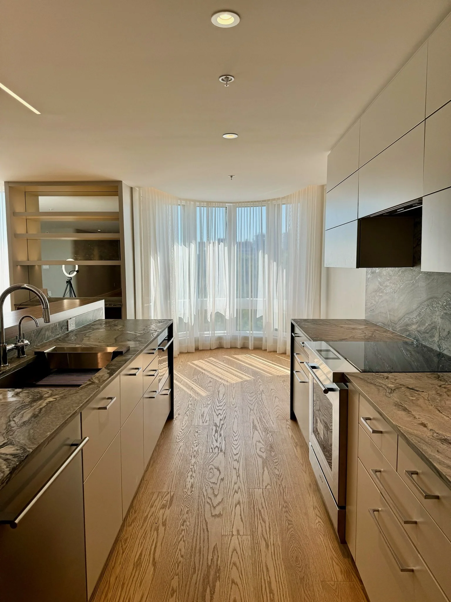 Modern kitchen with marble countertops, beige cabinets, wooden flooring, large window with sheer curtains, and ceiling lighting.