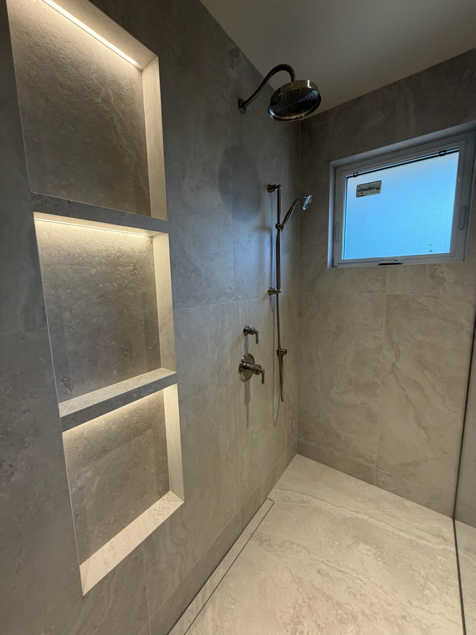 Shower area with two shower heads and a window, tiled walls and floor.