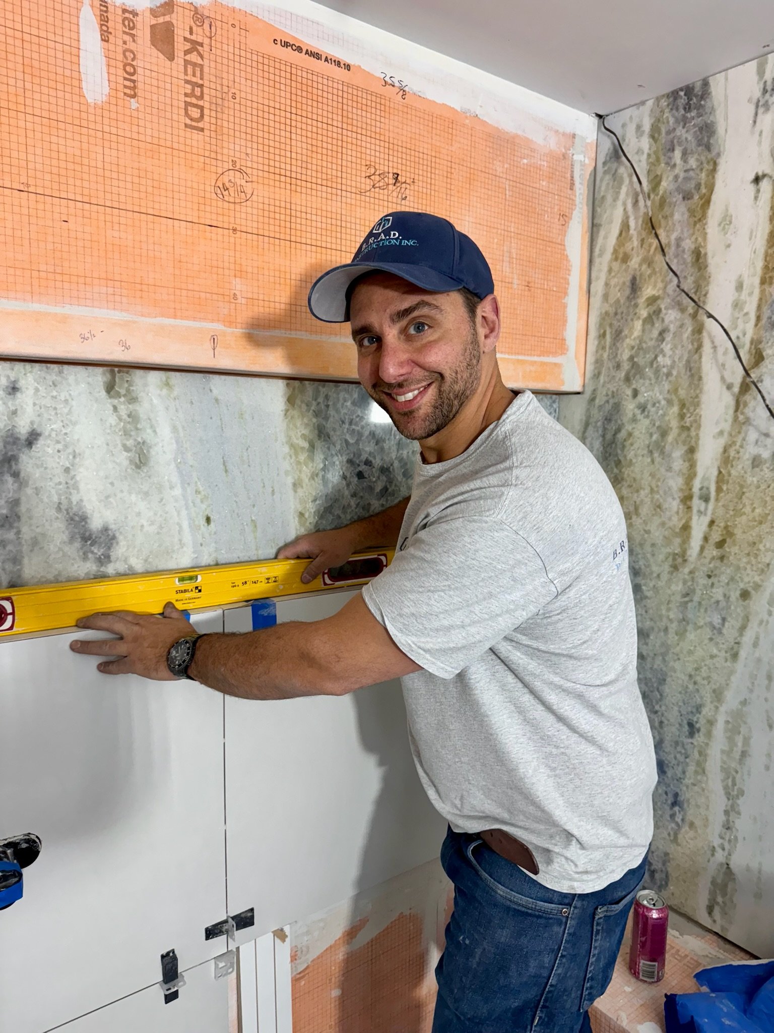 A man wearing a blue cap and gray t-shirt is using a level to align white cabinets during construction. He is smiling and working in a room with unfinished tile and insulation on the walls.