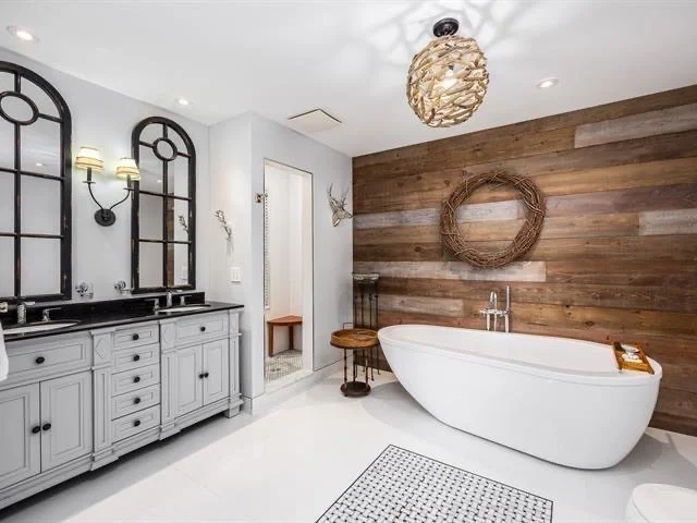 A modern bathroom with a white freestanding bathtub against a wood-paneled wall, a gray double vanity with black fixtures and mirrors, a decorative wreath on the wall, and a patterned rug on the white floor.