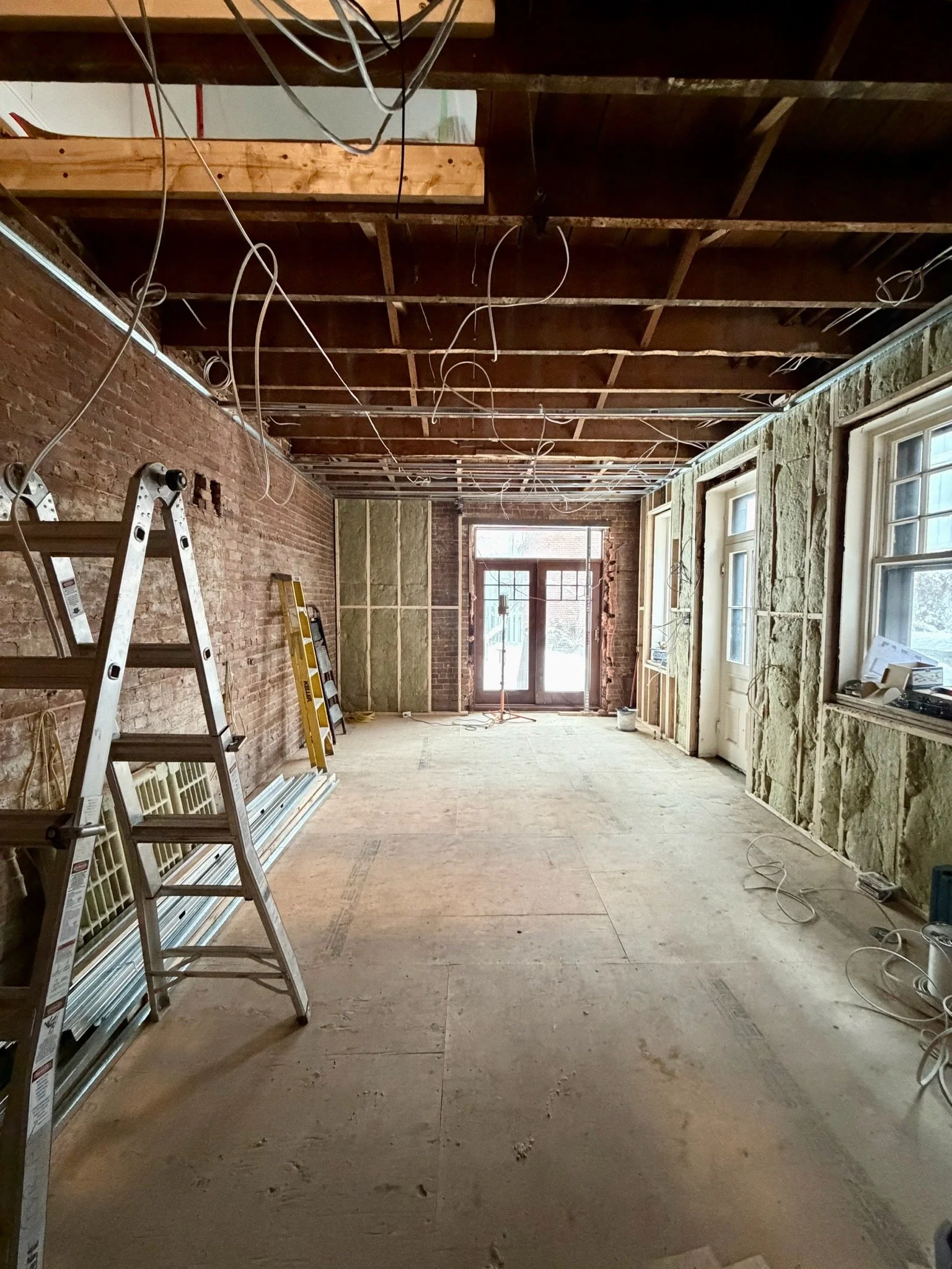 Interior of a room under construction with exposed brick wall, electrical wiring hanging from the ceiling, and windowed door at the end. Ladders and construction materials are present.