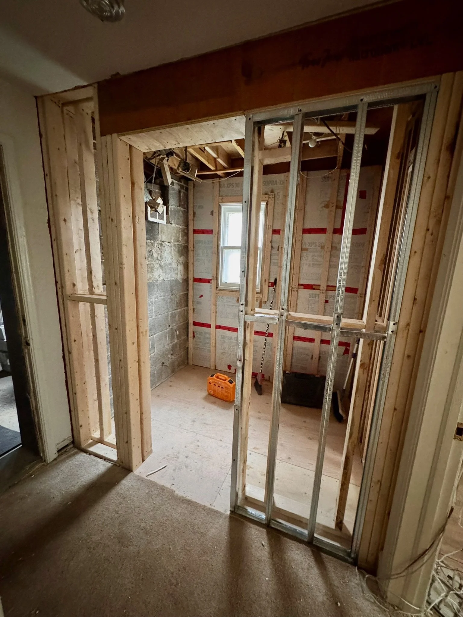 Interior of a house under construction with exposed wooden framing, a small window, and construction tools on the floor.