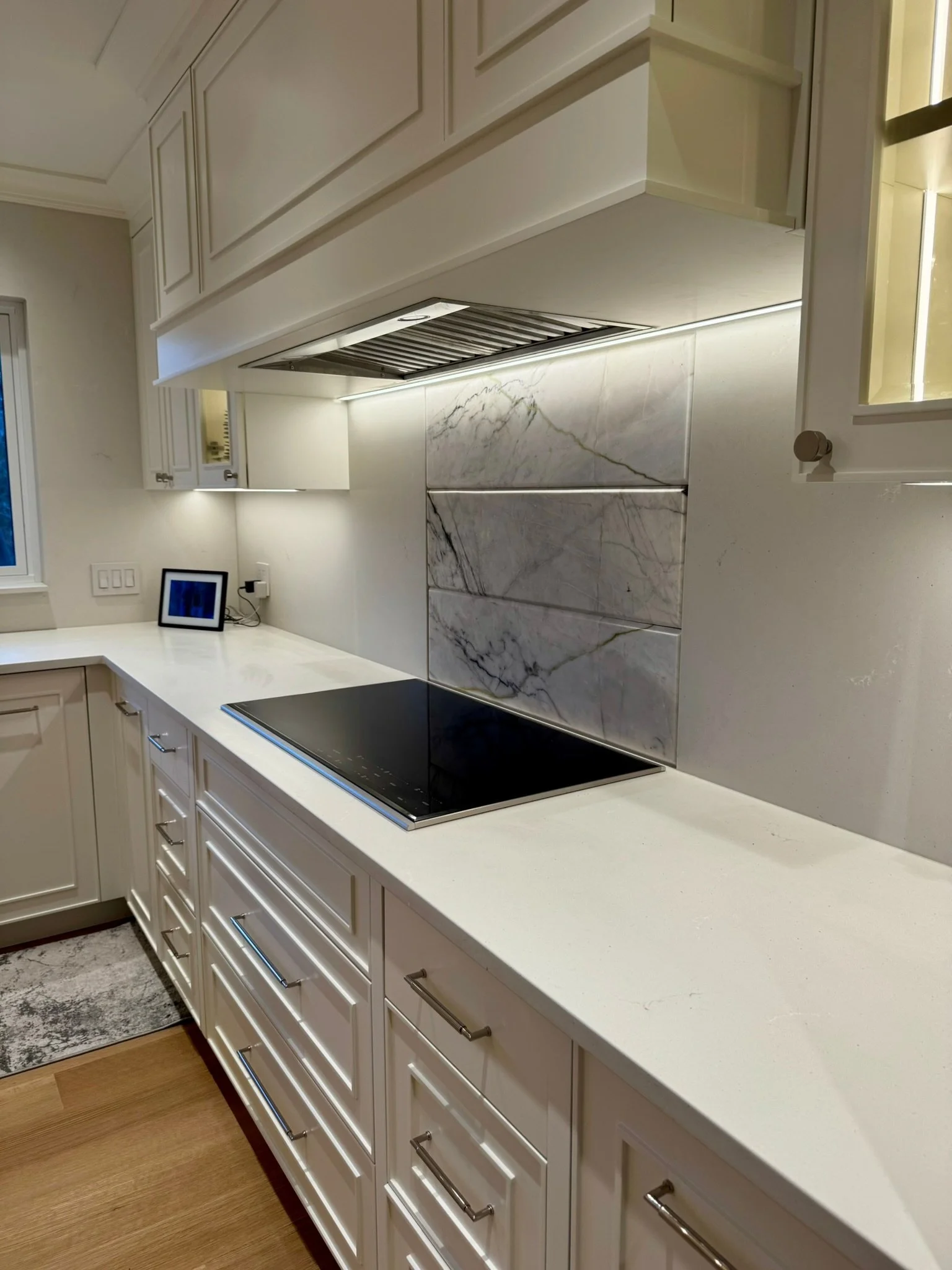 View of a modern kitchen with white cabinetry, marble backsplash, and a black cooktop on a white countertop. A small framed picture and some outlets are visible on the wall.