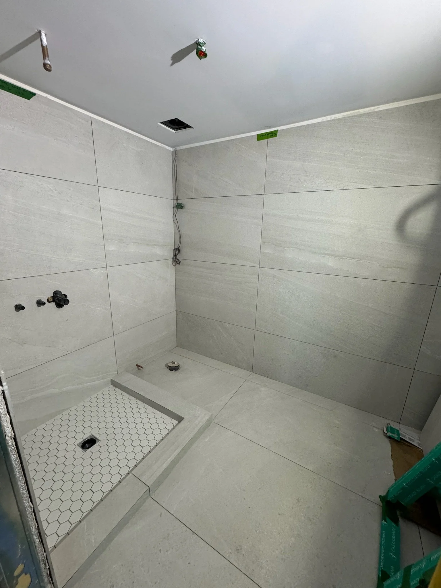 A bathroom under renovation with tiled walls and floor. There are plumbing fixtures, electrical wires, and an unfinished shower area.