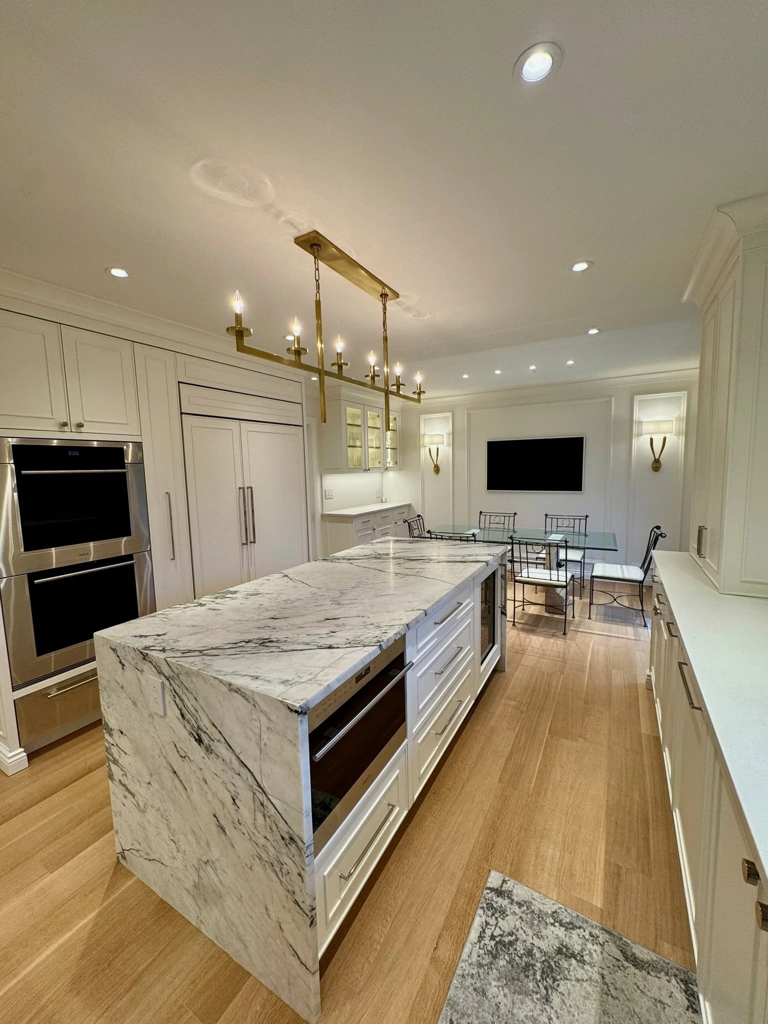 Modern kitchen with white cabinets, marble island, wooden flooring, and stainless steel appliances. There is a dining area with a glass table and black chairs, wall sconces, a large flat-screen TV, and a chandelier hanging from the ceiling.