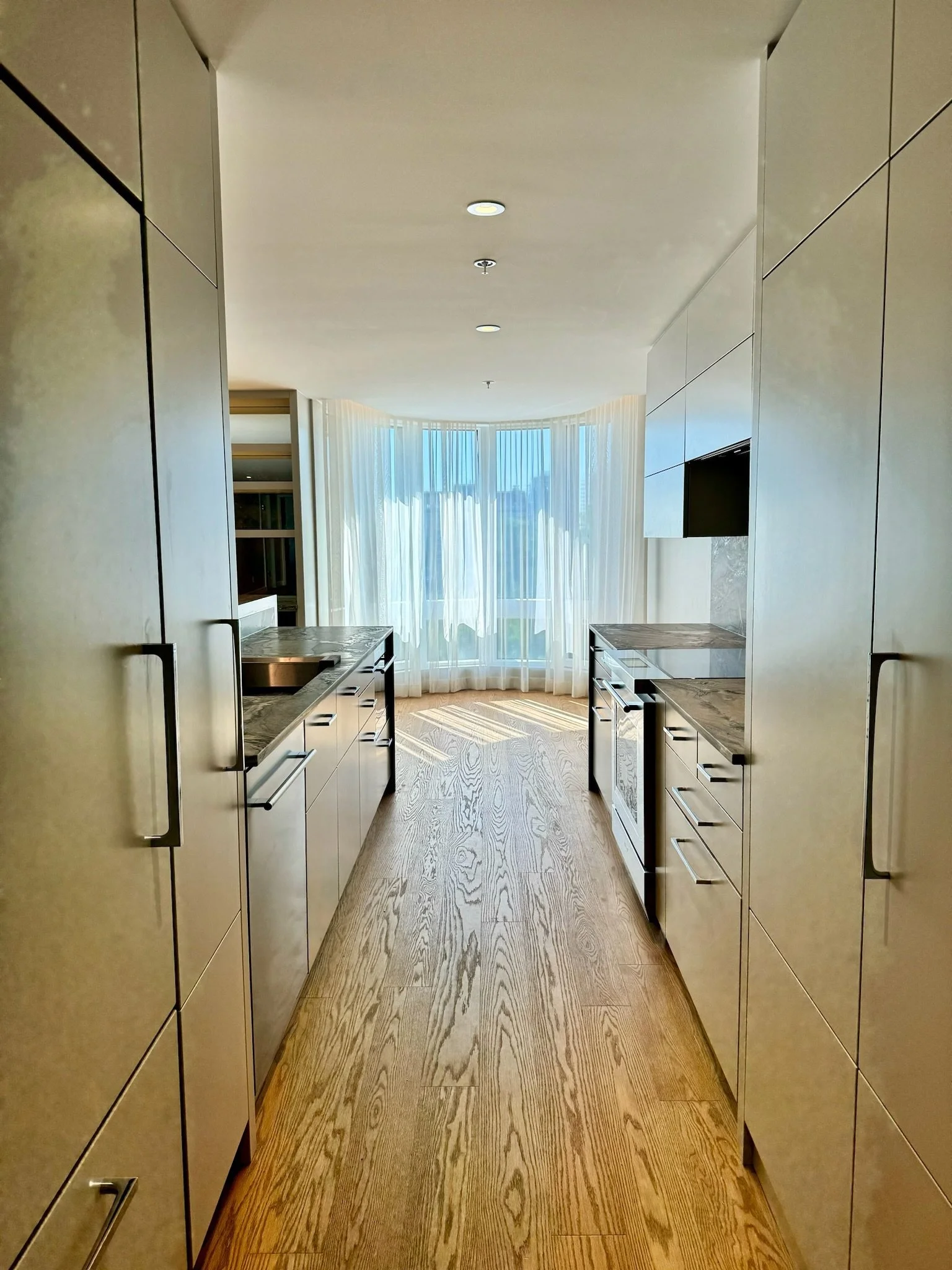 A modern kitchen corridor with beige cabinets on both sides, wooden flooring, and large windows with sheer white curtains letting in natural light.