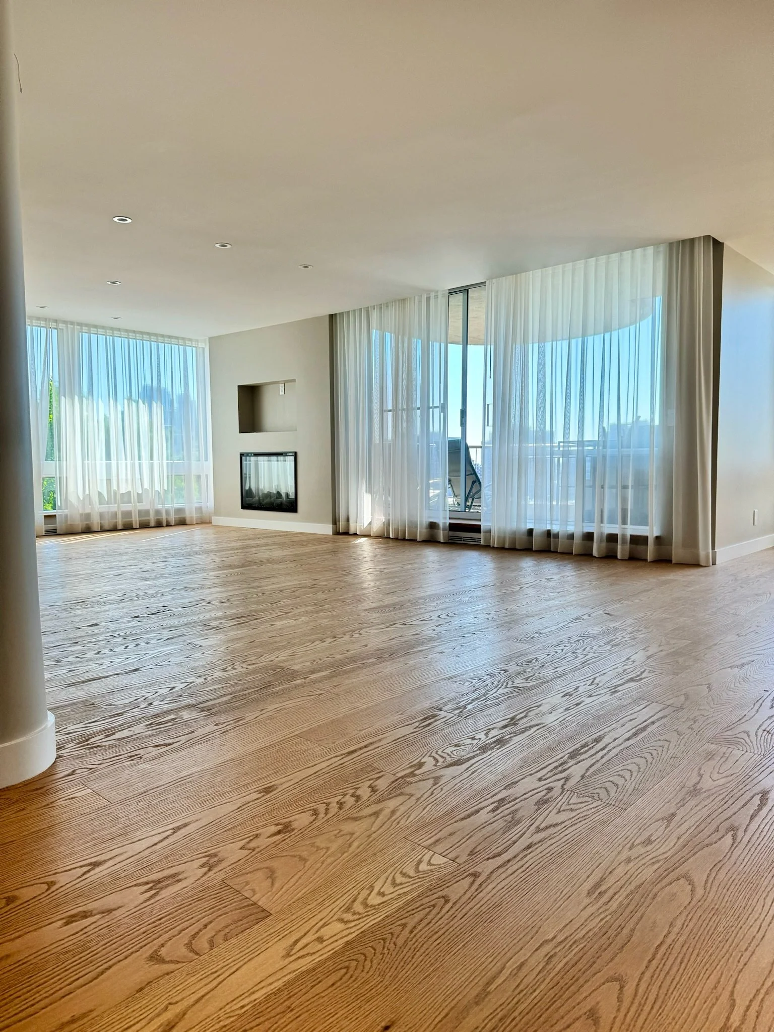 Living room with light wood flooring, large windows with sheer curtains, and a wall-mounted fireplace.
