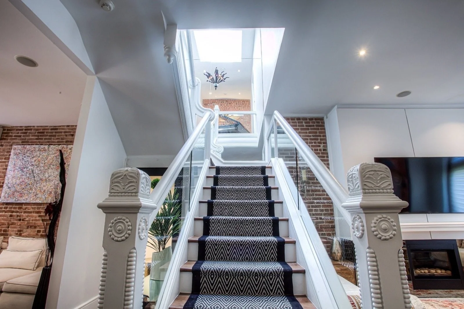 Indoor staircase with a black and white runner rug, white railings, and a glass wall on one side, leading to an upper floor with a skylight and decorative plant hanging chandelier.